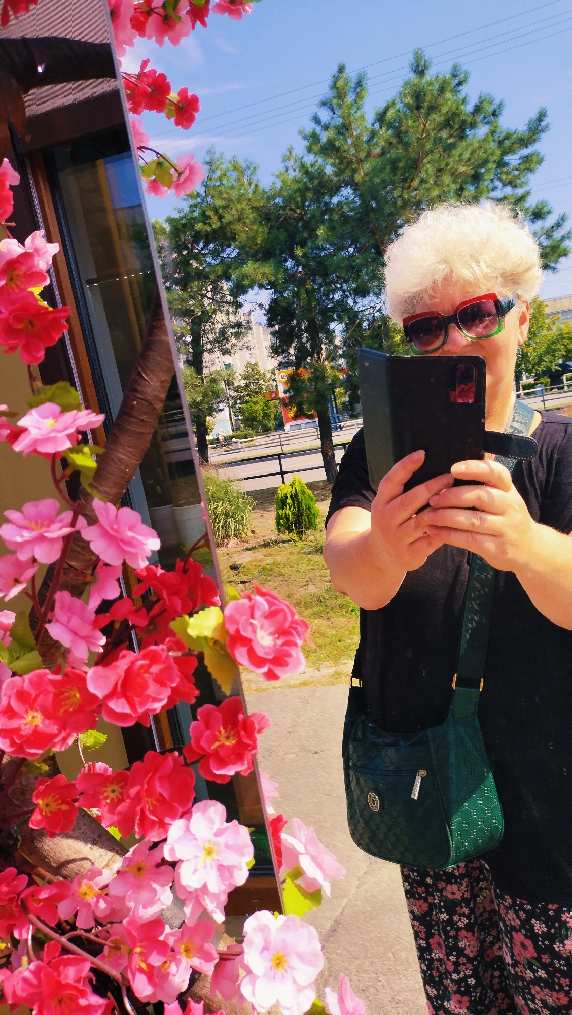 A woman with a white curly hairstyle and large, colorful sunglasses takes a mirror selfie outdoors. She is wearing a black t-shirt and a floral skirt, and a dark green patterned crossbody bag is slung over her shoulder. The background shows greenery, trees, and a street with power lines overhead. The scene is set in the city of Horishni Plavni, Ukraine, evident from the reflection in the mirror which shows a building and signage characteristic of the region. The mirror itself is positioned to reflect a cluster of artificial pink and red flowers in the foreground.