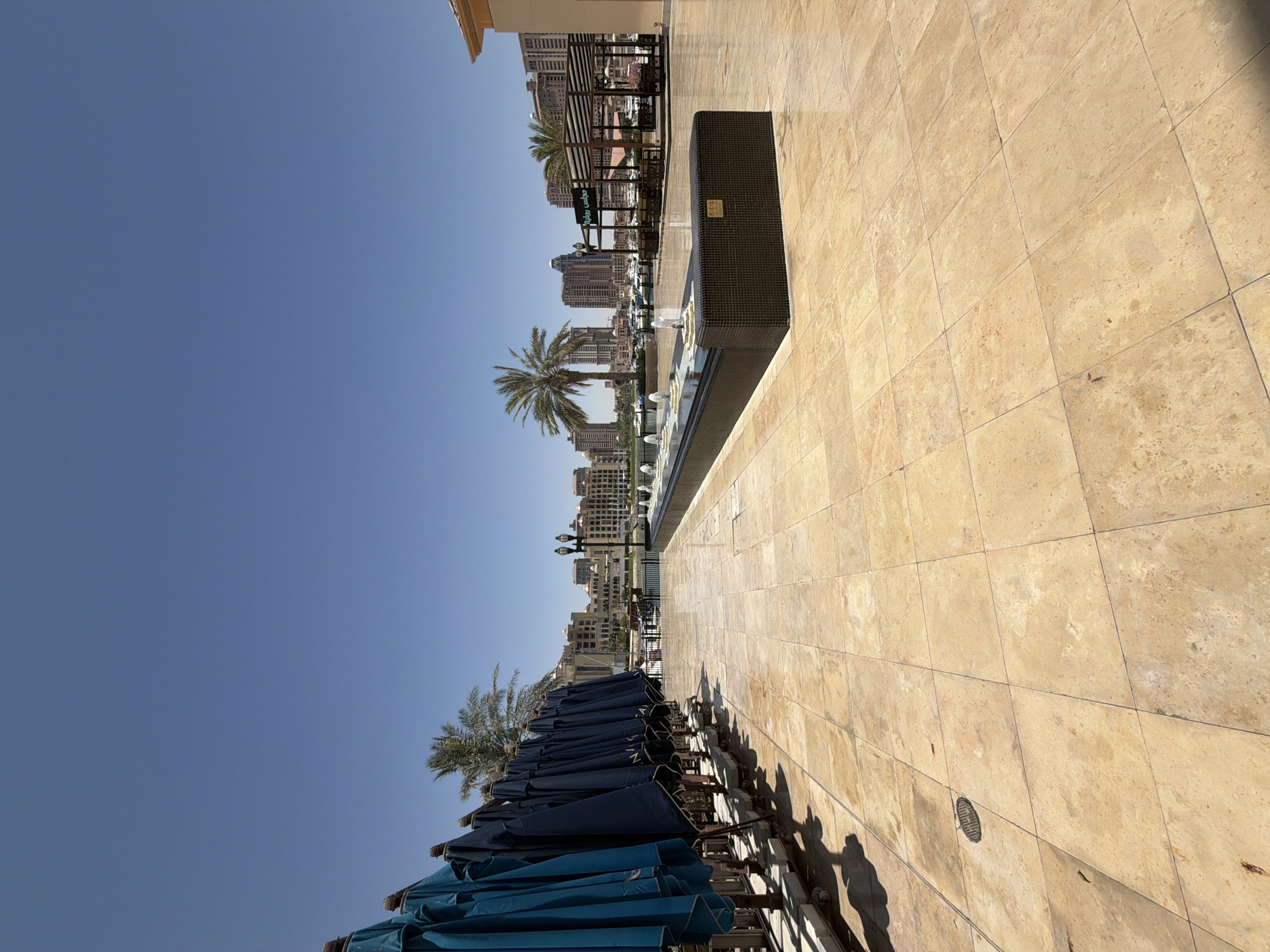 In a sunny outdoor setting, a row of closed navy blue and turquoise umbrellas are lined up on the left side of a spacious, beige tiled patio. A rectangular, dark tiled fountain with multiple jets of water is centrally located in the mid-ground. In the background, a city skyline with modern architecture and palm trees is visible under a clear blue sky. Several boats are moored in the water in front of the buildings. To the right, a covered seating area with tables and chairs is set up, and a sign with Arabic writing is displayed. The overall scene suggests a public leisure area, possibly a waterfront promenade or plaza.