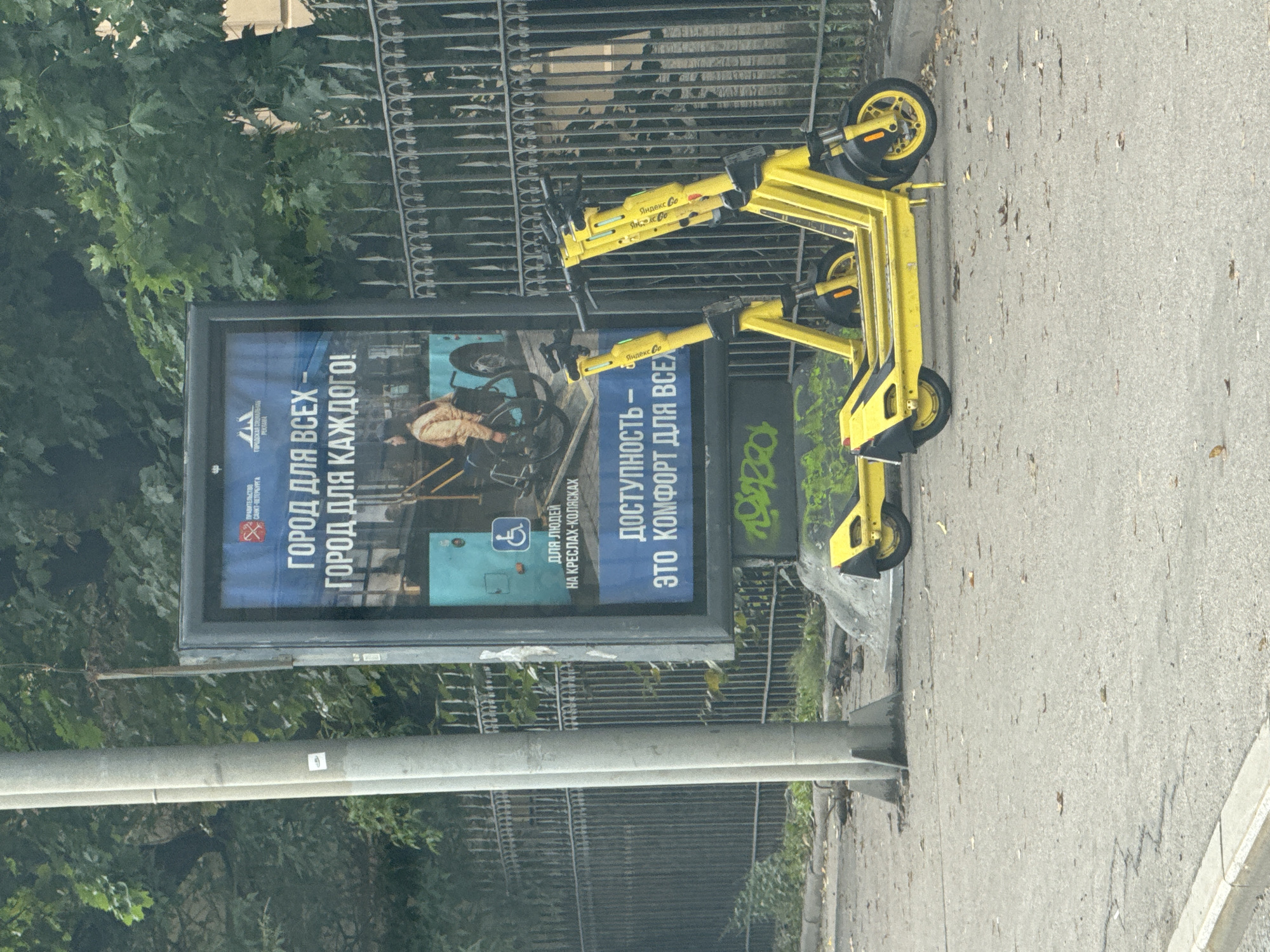 In an outdoor urban setting, likely in Saint Petersburg, Russia, a public advertising billboard stands prominently, displaying a message in Russian. The billboard, with a blue background, features text that translates to "A CITY FOR ALL - A CITY FOR EVERYONE!" and highlights "ACCESSIBILITY - IT'S COMFORT FOR ALL." An image on the billboard depicts a person in a wheelchair near a bus, emphasizing accessibility for people with disabilities. Two bright yellow electric scooters are parked in the foreground, leaning against a decorative metal fence. The scooters appear to be part of a shared service, as evidenced by branding on their handlebars. The scene is set on a paved sidewalk next to a street, with trees and foliage visible in the background, suggesting a park or green area adjacent to the urban environment. The overall impression is a city promoting inclusivity and modern transportation options.