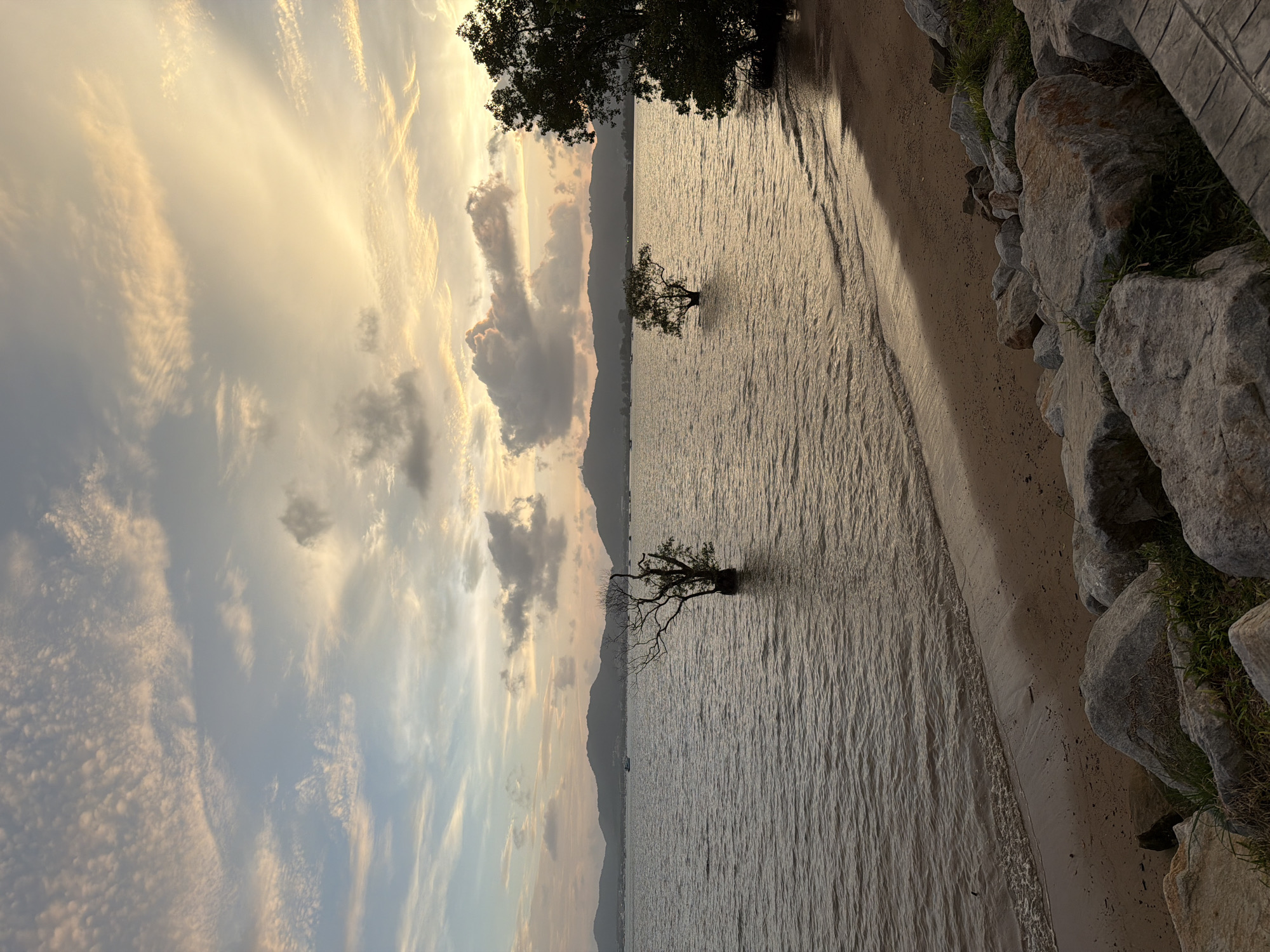 A tranquil sunset scene unfolds over a calm body of water in Wichit, Thailand. The sky is painted with soft hues of orange, yellow, and blue, with scattered clouds casting delicate shadows. In the foreground, a sandy beach is bordered by large, grey rocks and lush green grass. The water gently laps at the shore, reflecting the warm light of the setting sun. Two solitary trees stand partially submerged in the water, their bare branches reaching towards the sky. In the distance, a line of mountains forms a majestic backdrop, with a small boat visible on the water. The overall atmosphere is one of peace and serenity.