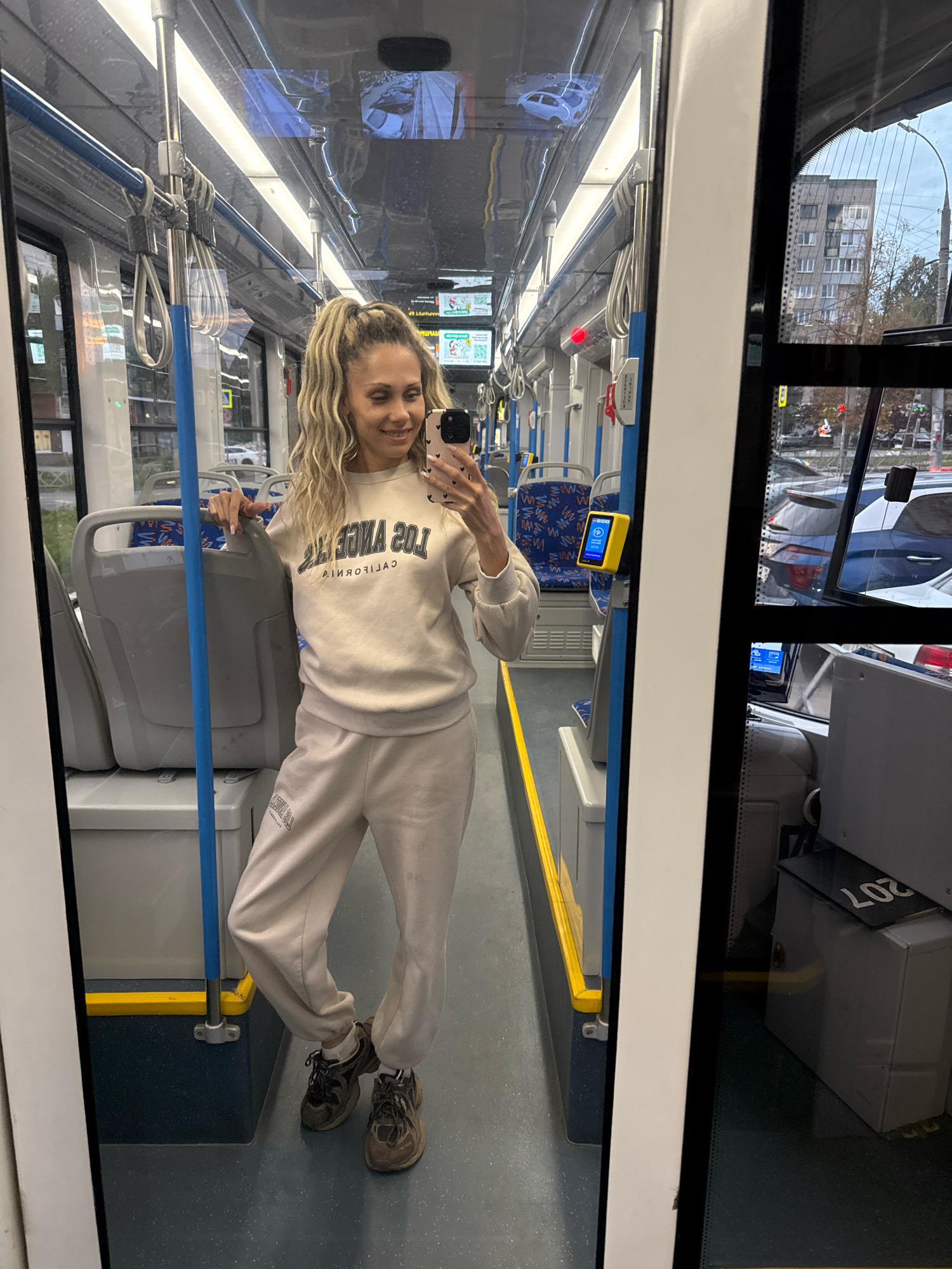 In Rusanovo, Russia, a woman with blonde, curly hair tied in a high ponytail stands inside a tram. She is wearing a beige sweatshirt with "LOS ANGELES CALIFORNIA" printed on it and matching beige sweatpants. She holds a phone in her right hand, taking a selfie in the tram's mirror. The tram interior features gray seats with blue and red patterns, blue poles, and yellow safety markings on the floor. The reflection in the mirror shows the tram's interior, including lights and windows. Outside the tram, visible through the windows, are buildings and parked cars.
