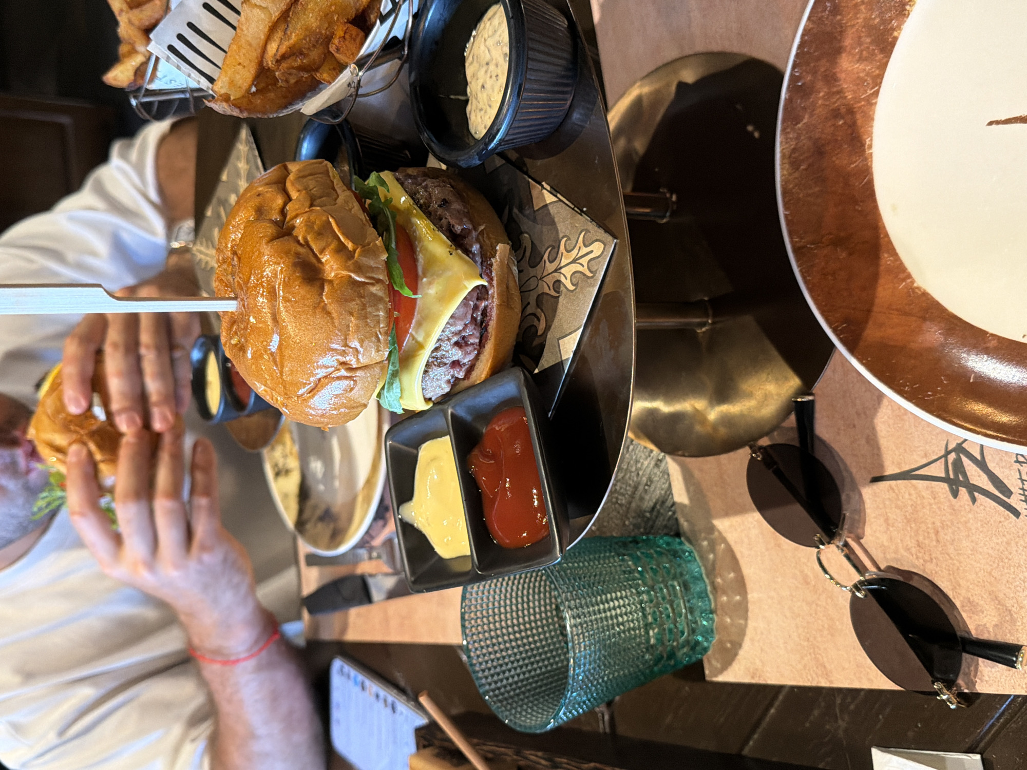A photograph taken in Choeng Thale, Thailand, shows a person at a restaurant table with a burger and fries. The burger is skewered and topped with cheese, tomato, and lettuce. There is a side of white sauce and a dish of ketchup and mustard next to the burger. The table is set with a plate and a glass of water. There are sunglasses on the table. The person is holding another burger in their left hand, with a red bracelet on the wrist.