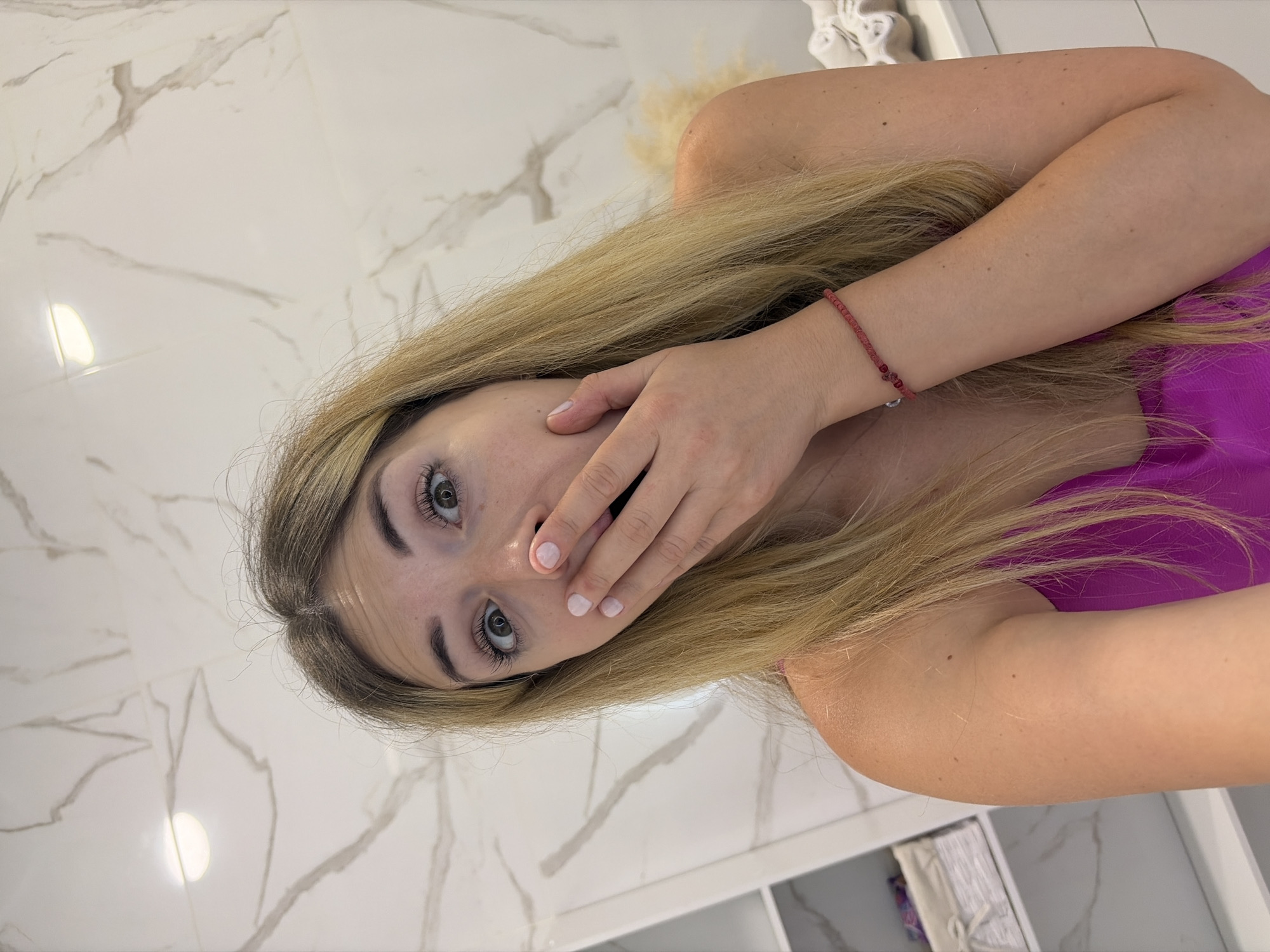 A young woman with blonde hair and green eyes is captured in a selfie. She is wearing a bright pink top and a thin red beaded bracelet on her right wrist. Her right hand is covering her mouth, and her eyes are wide with an expression of surprise. The background appears to be a modern bathroom with white marble walls and recessed lighting. A shelf with storage baskets is visible on the left side of the image. The scene is likely indoors, possibly in a home or hotel bathroom, in Budva, Montenegro.