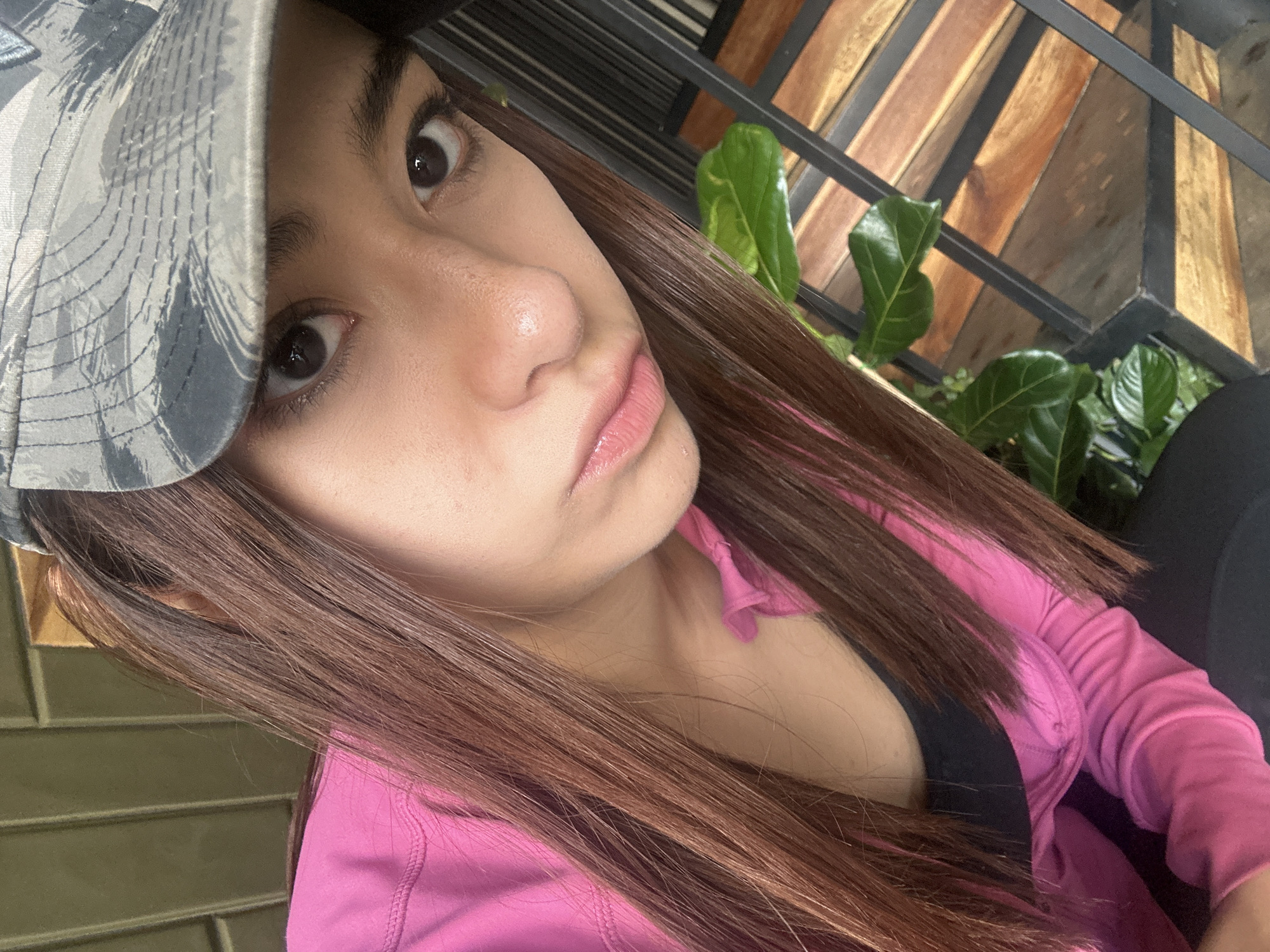 A close-up, angled portrait shows a young woman with light skin, dark brown hair, and brown eyes, wearing a camouflage baseball cap tilted to the side, a pink long-sleeved top over a black underlayer. She is positioned in front of an outdoor setting featuring a wooden and metal structure, with green foliage visible behind it. This suggests a location in a public space, likely near a building or garden. Based on the architectural style and vegetation, this is likely somewhere in Puebla, Mexico.