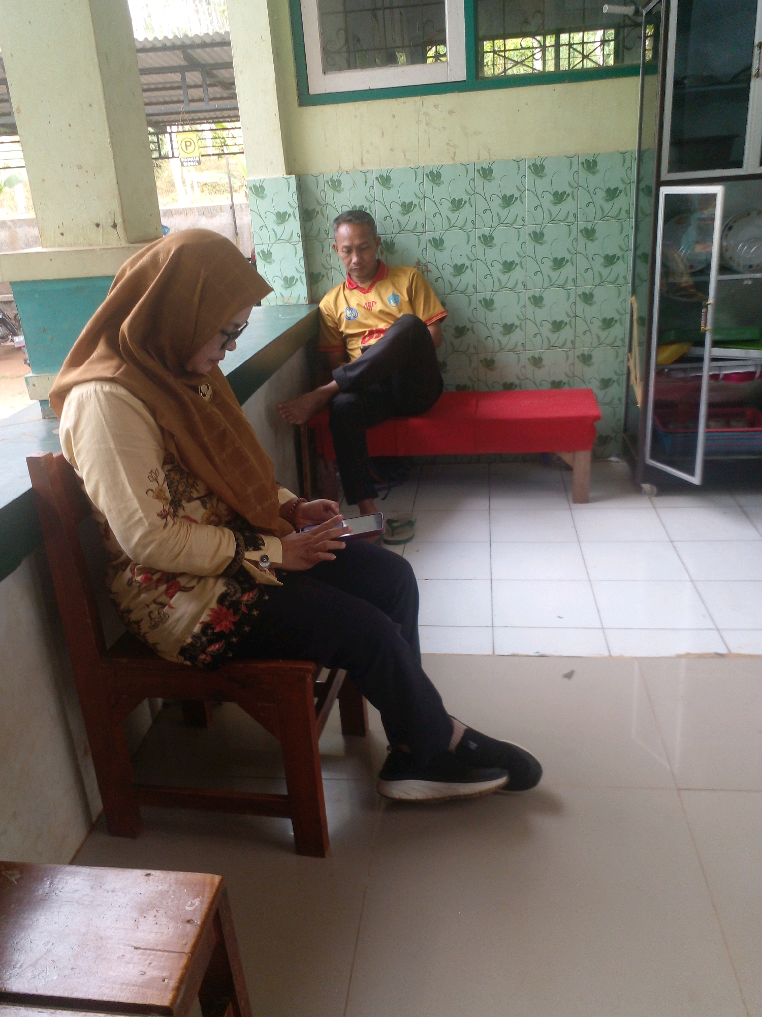 In an outdoor setting with a tiled floor and a light-colored wall, a woman wearing a brown hijab and glasses is seated on a wooden chair, looking down at a smartphone in her hands. She is dressed in a long-sleeved cream-colored shirt with floral patterns and dark pants. To her right, a man in a yellow shirt and dark pants is seated on a red bench, facing away from the camera. In the background, a green patterned wall is visible, along with a white cabinet with glass doors. A portion of a covered structure with a metal roof is visible in the upper left. The scene appears to be located in Banyuputih, Indonesia.