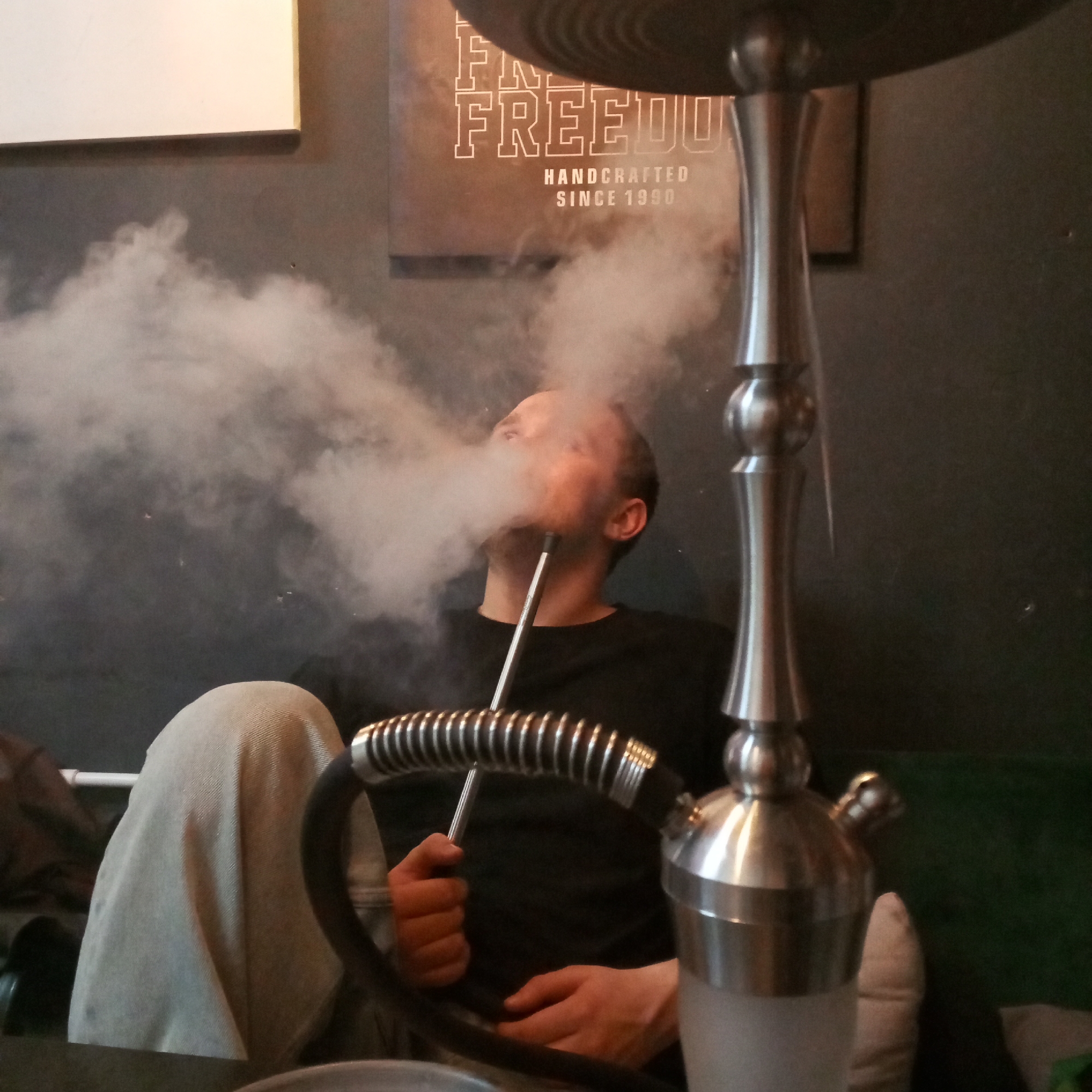 A man is sitting down while smoking a hookah indoors. He is wearing a black shirt and light-colored pants. The hookah is silver and black, with a hose in his hand. Large clouds of smoke surround his head. Behind him is a dark wall with two framed pictures. The text on one frame reads "FREEDOM HANDCRAFTED SINCE 1990." The location is identified in the prompt as Chaikovsky, Russia.