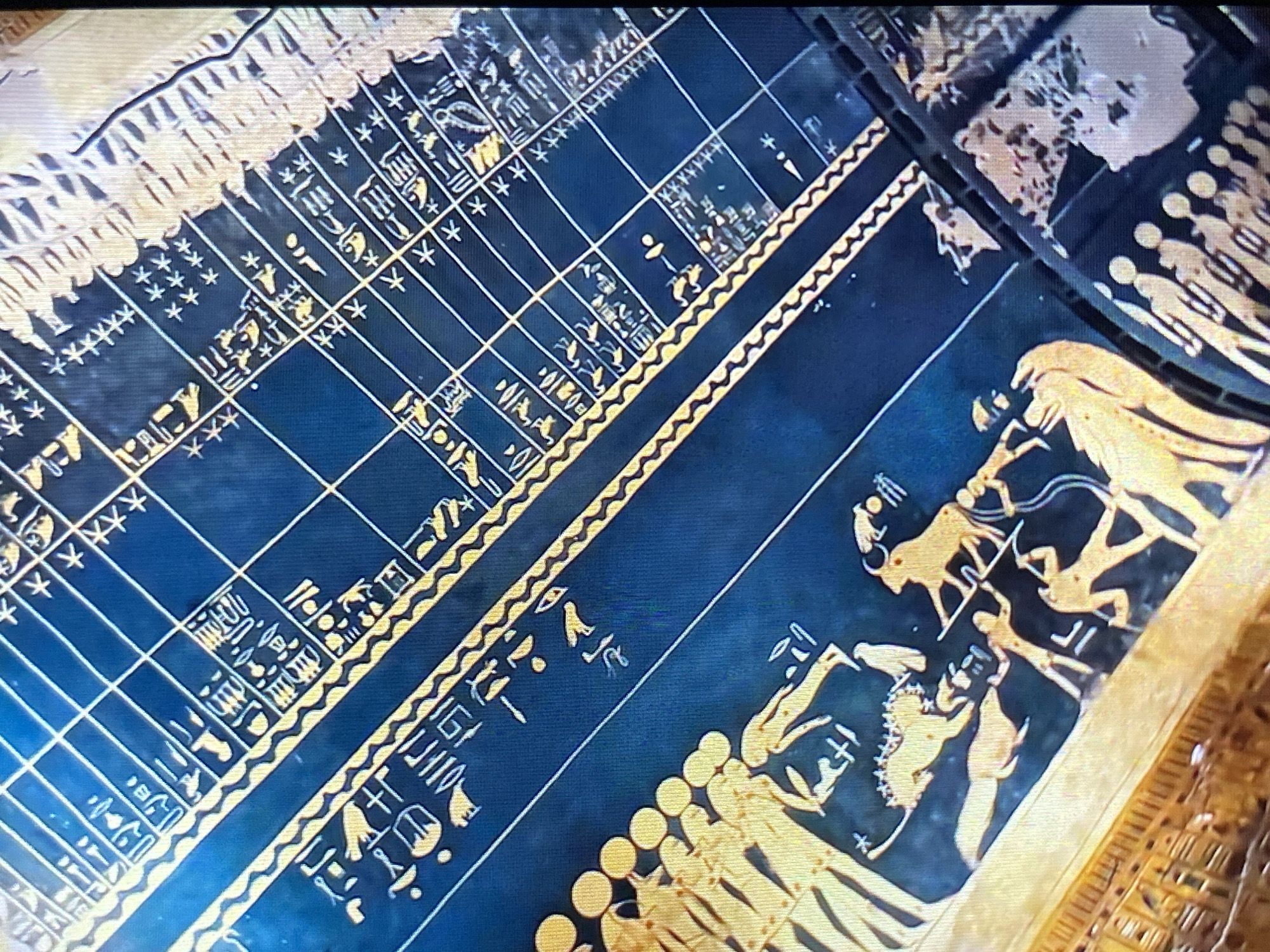 The image shows a detailed illustration, seemingly from an ancient artifact. The background is dark blue, overlaid with golden yellow drawings.
On the left side, there is a grid pattern with numerous hieroglyphic-like symbols and star-like shapes filling each cell. These symbols are rendered in golden yellow.
To the right of the grid, a decorative border consisting of a wavy line pattern separates the grid from a series of illustrations. The illustrations appear to depict scenes involving humans and animals, possibly in a mythological context. The figures are also in golden yellow against the dark blue background. The style is reminiscent of ancient Greek or Egyptian art.
The location is not depicted within the image. Therefore, I cannot determine that it is in the city of Tinaquillo, Venezuela.