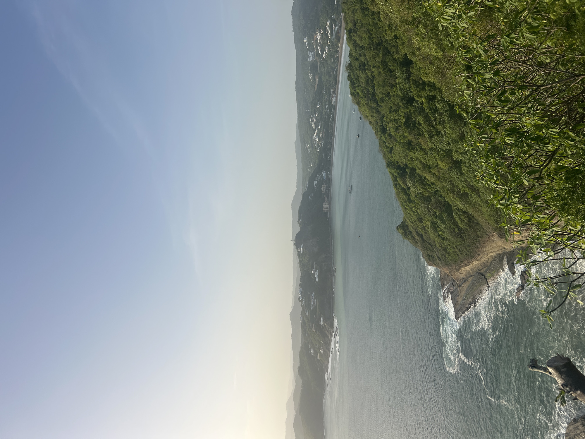 This image captures a scenic overlook of the bay in San Juan del Sur, Nicaragua. The foreground features lush green foliage and the edge of a cliff, with a weathered log in the lower left corner. Below the cliff, the turquoise ocean crashes against rocky shores, creating white foam. The middle ground showcases the expansive bay, dotted with several small boats. Along the coastline, white buildings are nestled amidst rolling, tree-covered hills, extending into the distance where layered mountains are visible under a pale, hazy sky. The overall scene suggests a tropical, coastal environment, likely captured during daylight hours.