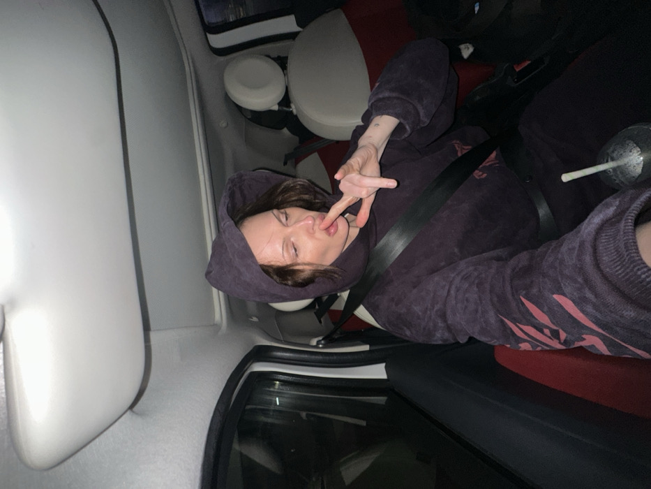 A person wearing a dark purple hoodie and pants, and a seatbelt is inside of a car. The person's left hand is near their mouth with their index and middle fingers extended. The car’s interior is mostly white, with red seats. A cup with a straw is visible. The image is taken from an off-kilter angle, with the car door in the foreground. The lighting is dim, suggesting it’s either nighttime or the inside of a darkened vehicle.