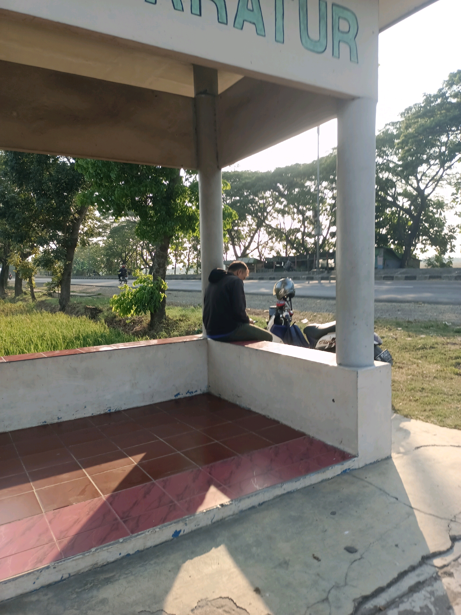 The image depicts an outdoor scene in what appears to be a public waiting area or shelter, possibly along a roadside. The structure is made of concrete pillars and a roof. The floor of the shelter is tiled with reddish-brown tiles. In the foreground, a person in a dark hooded jacket and green pants is seated on the edge of a concrete bench, facing away from the camera. Beside the person, a motorcycle with a silver helmet on its handlebars is parked. The background reveals a road with a few vehicles, trees, and grassy areas under a bright, sunny sky, suggesting it is daytime. The overall environment appears to be a somewhat rural or suburban setting within Subang, Indonesia. The sunlight casts shadows across the scene, indicating the time of day. There are some subtle signs of wear and tear on the structure, such as chipped paint on the lower parts of the walls.