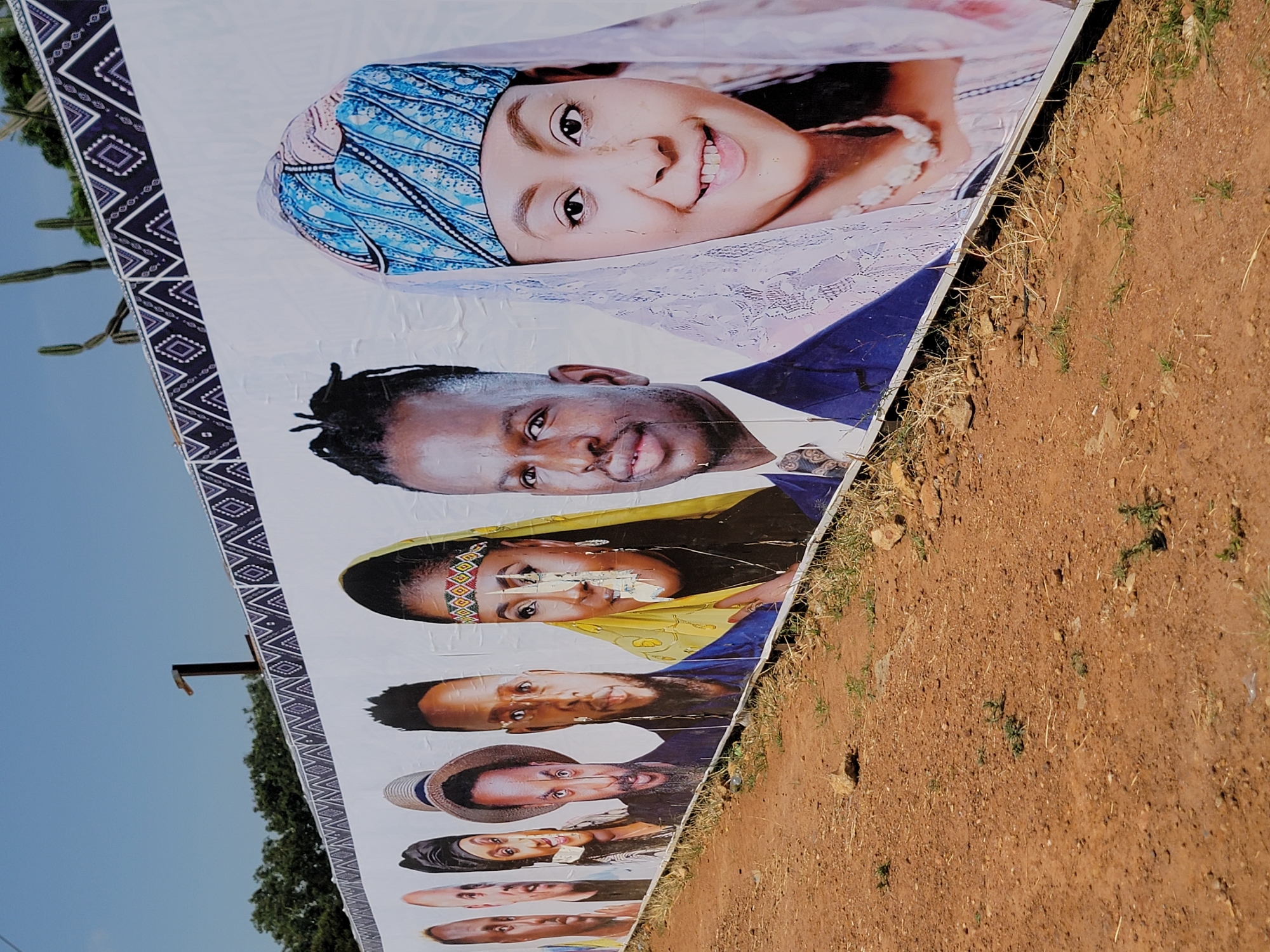 A photograph taken in Jos, Nigeria, shows a banner lying horizontally on reddish-brown earth. The banner features portraits of several people. The topmost portrait is of a young woman wearing a blue and white patterned head covering and a white, lace-like veil. Below her is a man in a blue suit. Subsequent portraits feature other individuals, including women in traditional headwraps and men wearing hats. The banner is bordered with a black and white geometric pattern. In the background, a blue sky and green trees are visible.