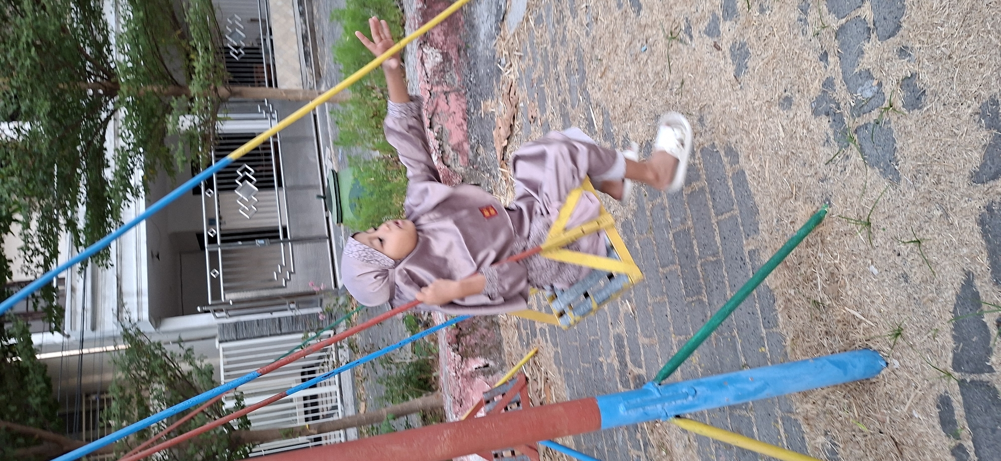 A young girl wearing a pink hijab and matching attire is on a swing set in a residential area that appears to be in Surabaya, Indonesia. The swing set is made of colored metal poles, with the seat being yellow. The girl is looking to her right, with her right arm extended forward. The ground around the swing set is a mix of brick paving and a light-colored mulch with sparse grass. In the background, a modern house with multiple levels and white siding is visible behind a metal fence. Trees with green foliage are present in the mid-ground and background. The overall lighting suggests it is daytime, possibly late afternoon.