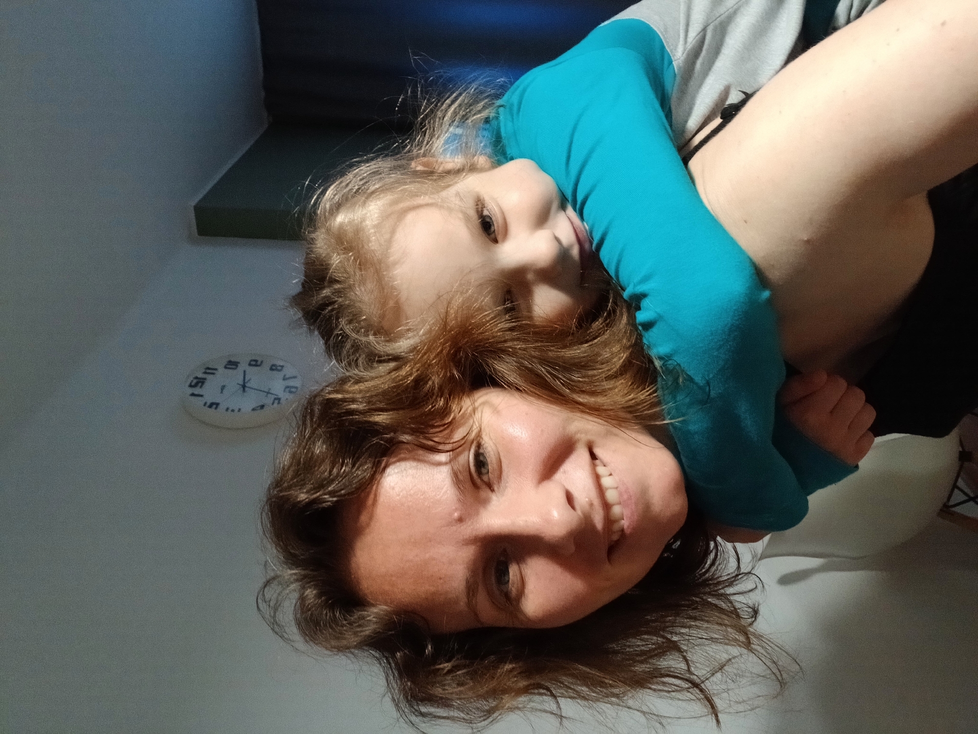 This is a close-up, selfie-style photograph of two individuals. On the left is an adult female with medium-length, wavy brown hair and a smiling expression. She appears to be looking directly at the camera. Embracing her from behind is a young child, likely a girl, with blonde hair. The child is wearing a turquoise long-sleeved shirt and is hugging the adult around the neck. The child is also smiling and looking towards the camera. The background is an indoor setting with a plain wall and a clock on the wall. The lighting suggests it is daytime. The image quality is typical of a smartphone camera, with some graininess. There is no specific information to determine the location within the city of Yekaterinburg, Russia.