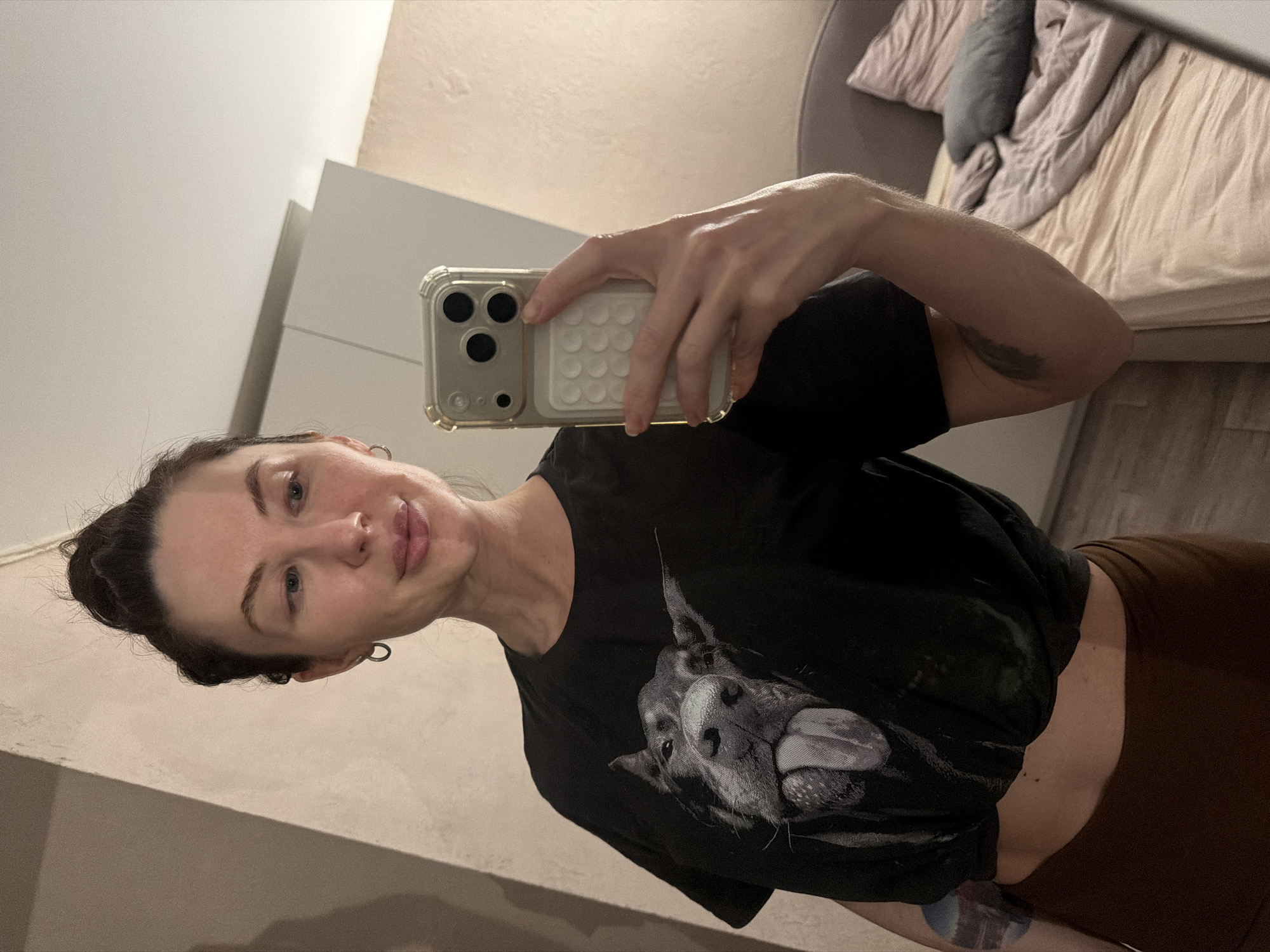 A person is taking a selfie in front of a mirror. The person has fair skin, dark hair pulled back in a bun, and is wearing a black t-shirt with a graphic of a dog on it and brown pants. They are holding a cell phone in their right hand. The phone has a light-colored case with a bumpy texture on the back. A glimpse of a room with a bed, bedding, and possibly a wardrobe is visible in the background. There is no indication this image was taken in Perm, Russia.