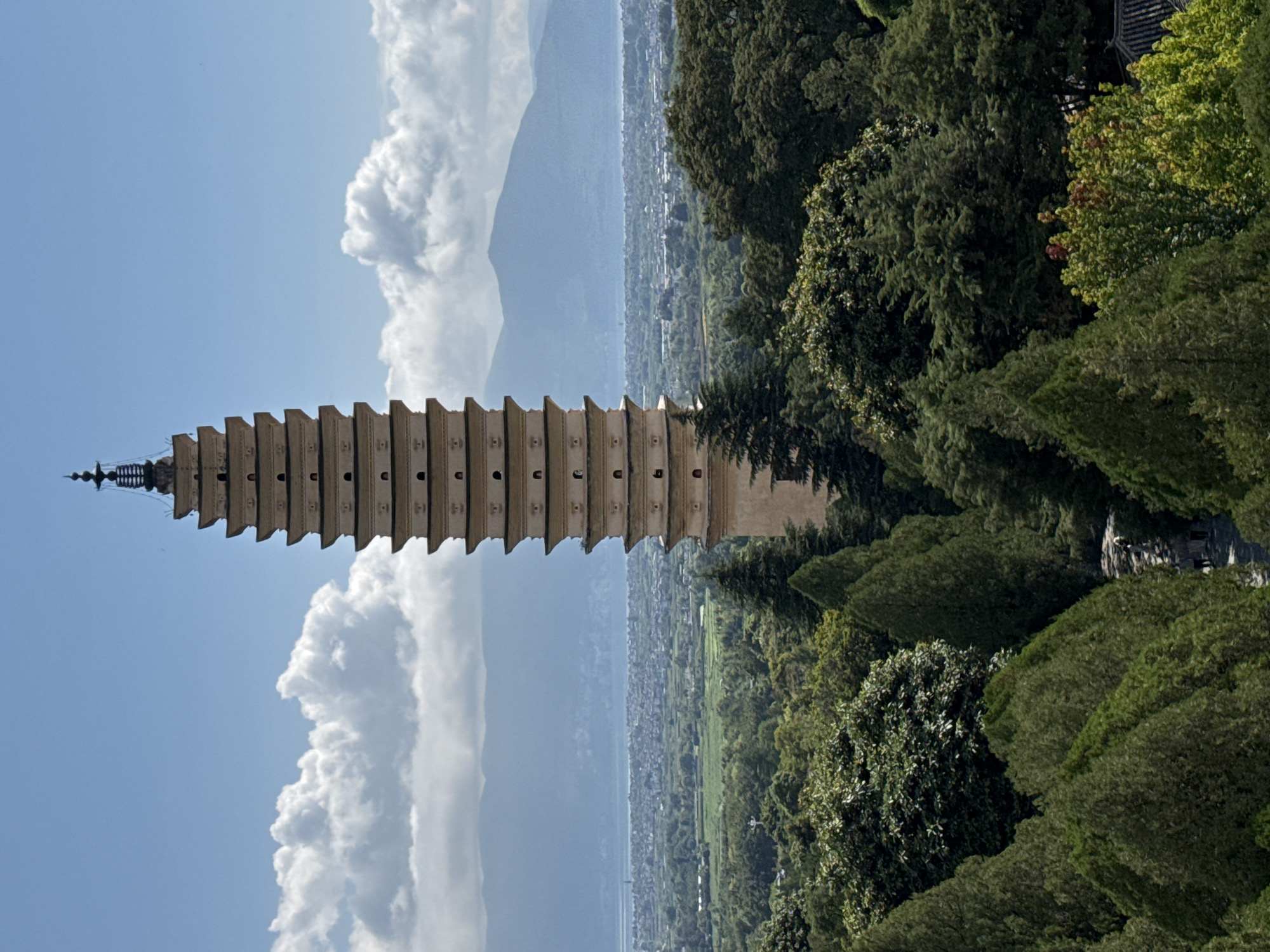 The image captures the Three Pagodas of Chongsheng Temple in Dali City, China, under a partly cloudy sky. The central pagoda, a tall, tiered structure with a dark, ornate finial, dominates the frame. It is surrounded by lush green trees, creating a natural foreground. In the background, a city stretches out towards distant mountains and a body of water, likely Erhai Lake. The scene is viewed from a slightly elevated position, looking down over a winding path with a few small figures visible, suggesting a tourist area or temple grounds. The architecture of the pagoda, with its distinct levels and decorative elements, is a prominent feature, set against a backdrop of natural and urban landscapes.