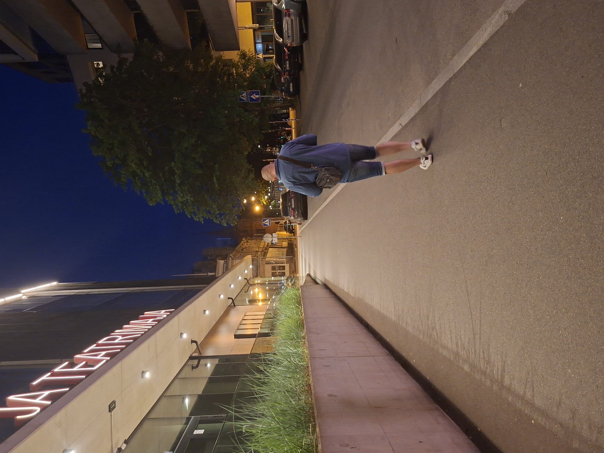 The image captures a street scene in Tallinn, Estonia, at dusk. In the foreground, a man with a shaved head is walking away from the camera on a paved sidewalk. He is wearing a blue denim shirt, denim shorts, white sneakers, and a brown shoulder bag. To his left, a modern building with large glass windows is visible, featuring illuminated red lettering that spells "JA TEATRIMÄJA." The building has a concrete planter box filled with tall green grass along its base. The street itself is lined with parked cars, and a large tree with dark green foliage stands on the right side of the road. The sky is a deep blue, indicating late evening. Streetlights and building lights illuminate the area.