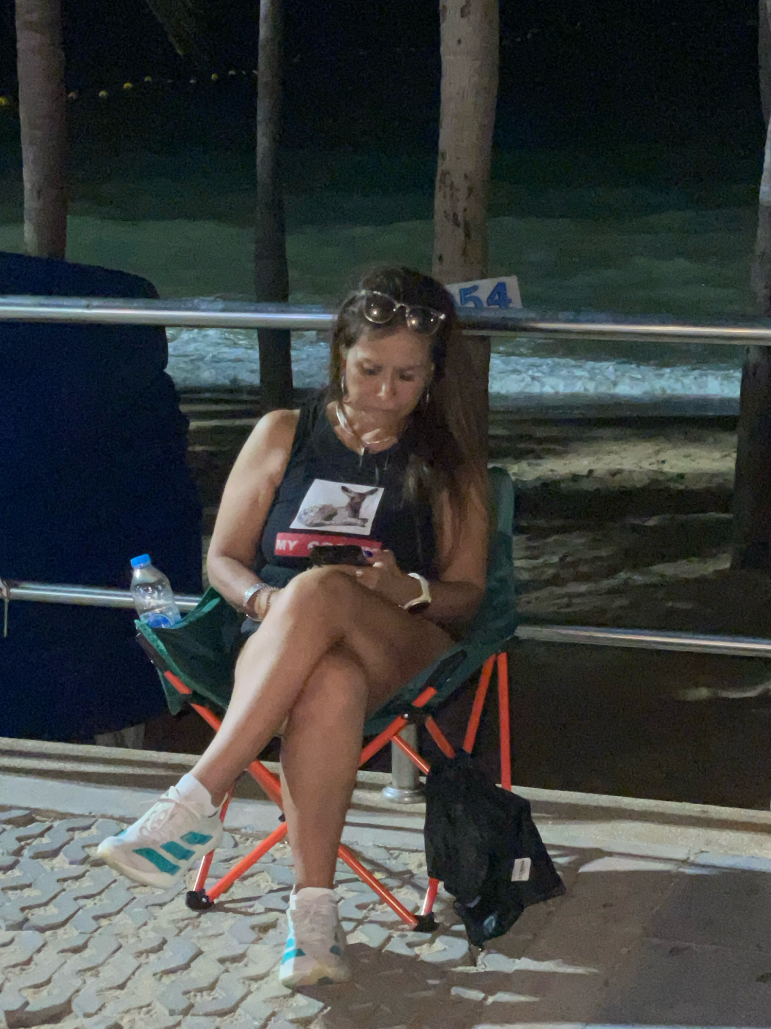 A woman is sitting in a foldable chair on a paved walkway near a beach at night. She is wearing a black tank top with a graphic of a deer and the word "MY" in red, denim shorts, white sneakers with teal accents, and sunglasses on her head. She is looking down at her phone. Next to her is a bottle of water and a black jacket is hanging on the chair. In the background, palm trees are visible, and the ocean and beach are dimly lit. A blue sign with the number "54" is mounted on a railing in the background.