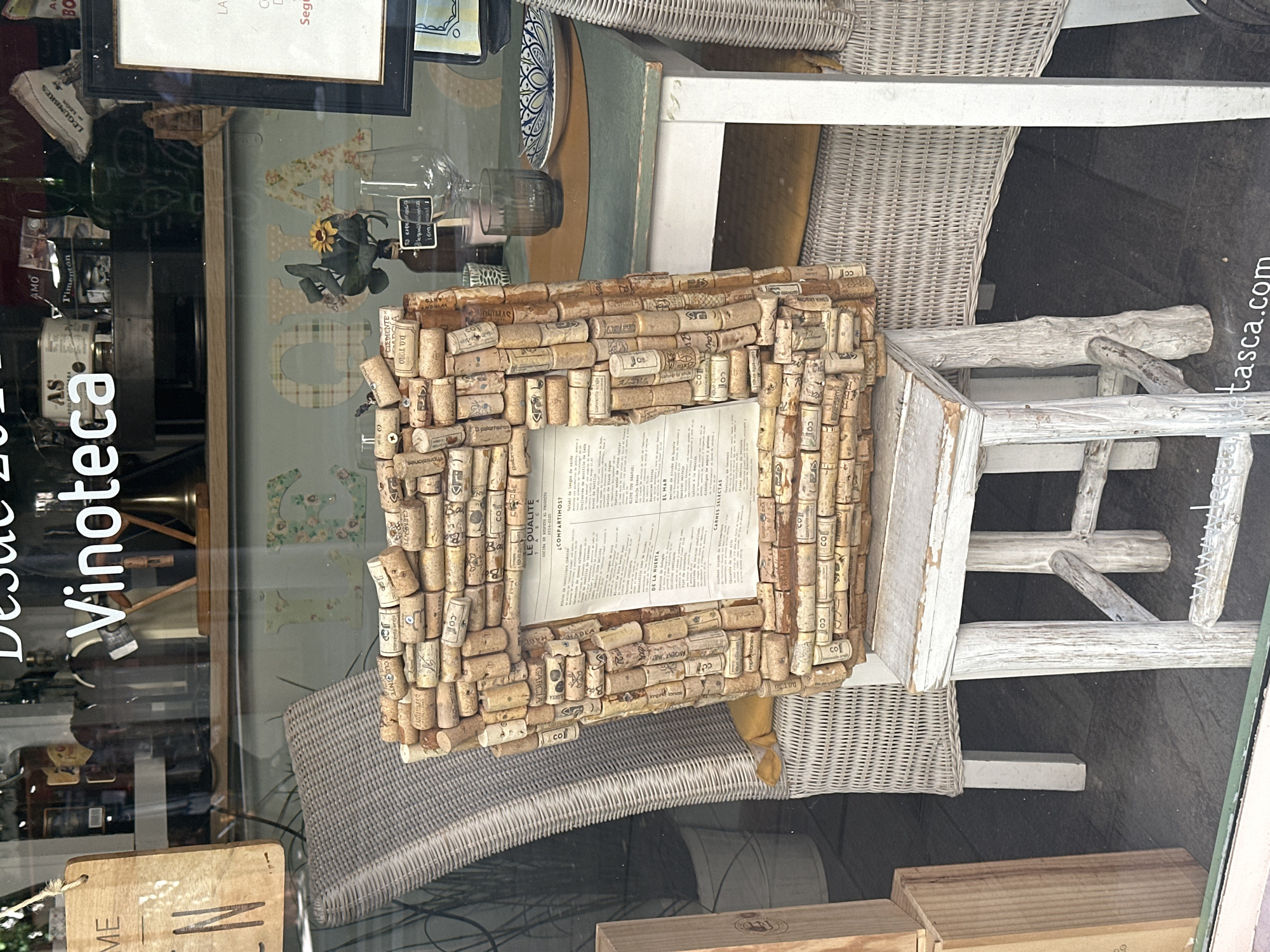 A detailed view of a restaurant window display in Madrid, Spain.
**Subjects and Objects:** The primary focus is a cork-framed menu or informational sheet positioned on a distressed white wooden stool. The frame is made of numerous wine corks, each bearing unique imprints and stamps. A sheet of paper is visible in the middle of the cork frame, which contains text. The background reveals elements of the restaurant interior, including wicker chairs, a framed sign hanging above, bottles, and other decorative items.
**Environment:** The display is inside a window, evidenced by the reflections visible on the glass surface. The store is called "Vintoteca Desde 2014". Text like "wetasca.com" is also printed to the right of the window.
**Action and Interactions:** There are no visible people or actions within the frame. The scene is a static display meant to attract customers or inform passersby.