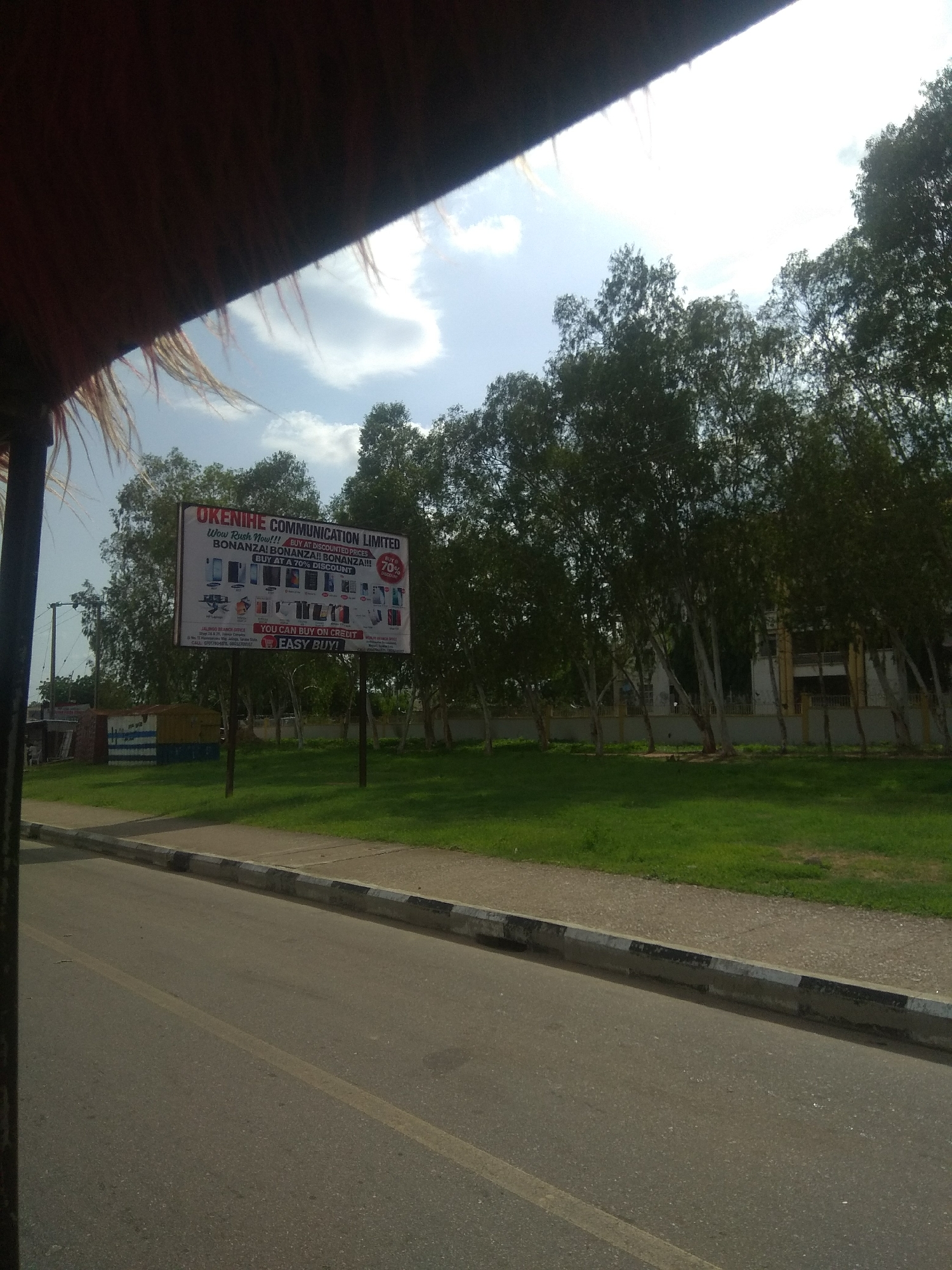 This is a medium shot, taken from a vehicle, of a billboard advertisement in Howayi, Nigeria. The billboard, positioned by the roadside, advertises "OKENIHE COMMUNICATION LIMITED" and promotes "BONANZA! BONANZA!! BONANZA!!!" with a 70% discount on purchases. It displays images of various mobile phones and electronic devices, suggesting a sale or promotion. The text also highlights the option to "BUY ON CREDIT" and states "EASY BUY!". Below the main advertisement, contact information for the "JALINGO BRANCH OFFICE" is visible, along with a phone number.
The scene is outdoors on a sunny day, with a bright blue sky and scattered clouds. Tall, slender trees with dense green foliage line the background, some leaning over the billboard. A neatly mown green lawn separates the road from the trees. The road itself is asphalt, with a single yellow line marking its center. The foreground shows the edge of the vehicle from which the photo is taken, with a dark brown, furry-like material obscuring the top left corner. The overall impression is a commercial advertisement set in a somewhat rural or suburban environment.