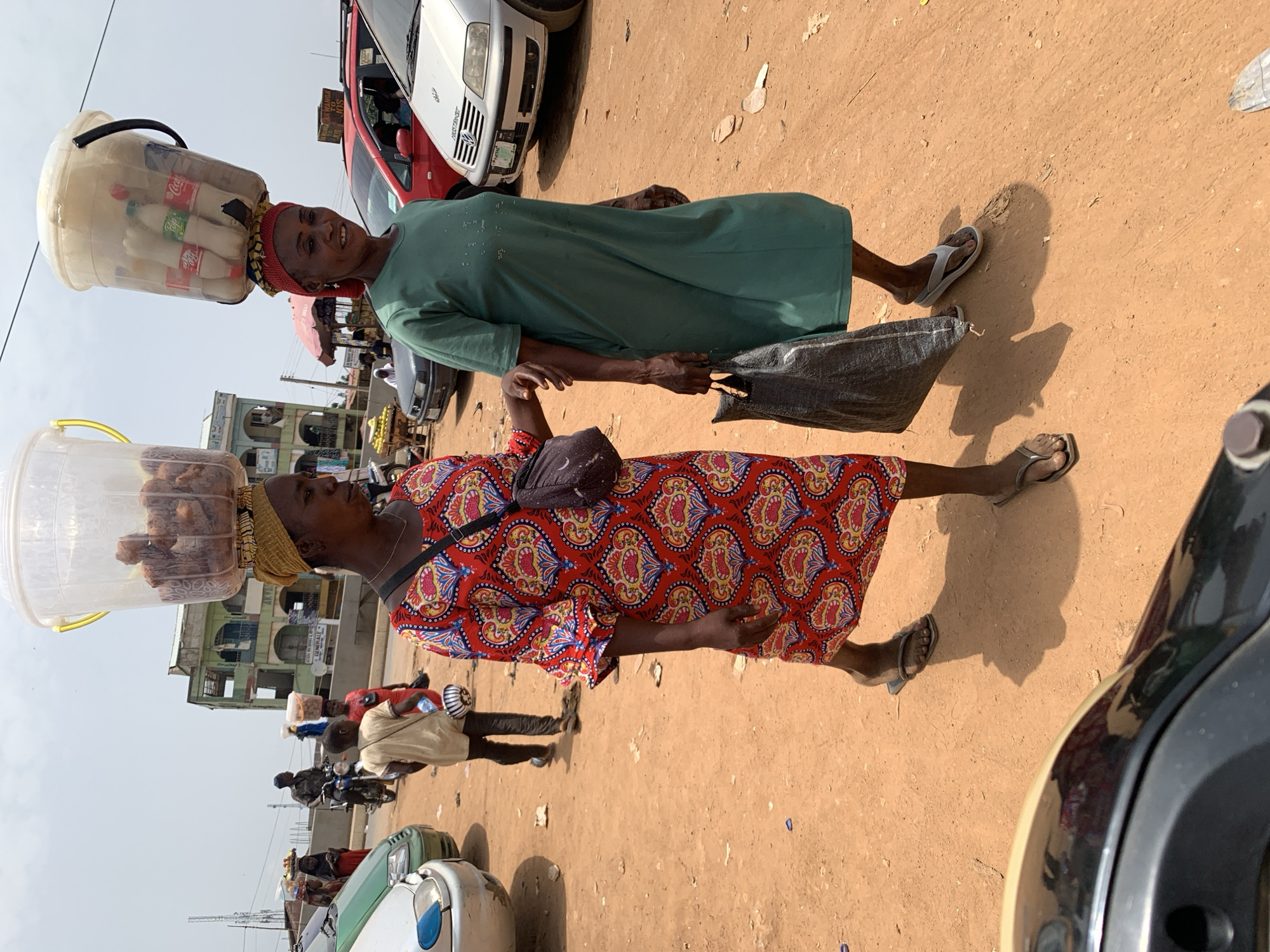 The image captures a scene in Akwanga, Nigeria, depicting two women walking along a dusty street. The woman closer to the camera is wearing a red patterned dress and has a container balanced on her head, filled with what appears to be fried food. She also has a small bag slung across her shoulder. The second woman is wearing a green dress and carries a gray bag. She has a container on her head filled with bottled beverages, seemingly Coca-Cola and Sprite. Cars are parked along the side of the road and buildings can be seen in the background, along with other people and motorcycles. The overall environment is urban, with evidence of commercial activity.