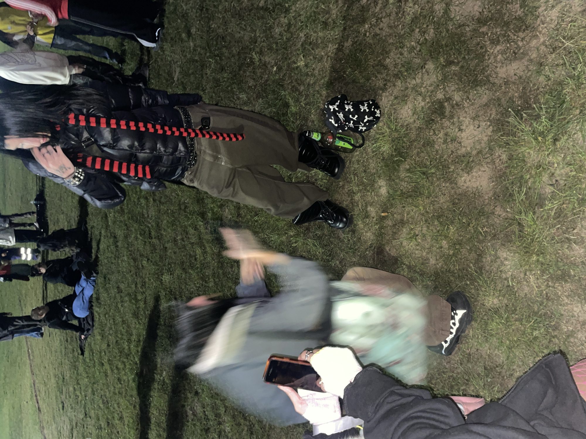 The image shows an outdoor scene at night. Two individuals are visible in the foreground, lying on the grass. One is wearing a black puffer jacket with a red and black striped accessory, green pants, and black boots. Next to them are a black bag with white skull patterns and two green bottles. The second individual is in motion, wearing a gray jacket. A person holding a phone is visible in the lower right corner. In the background, other people are standing or walking on the grass. The scene appears to be on a grassy field at night. No specific details indicate the location is in Buenos Aires, Argentina.