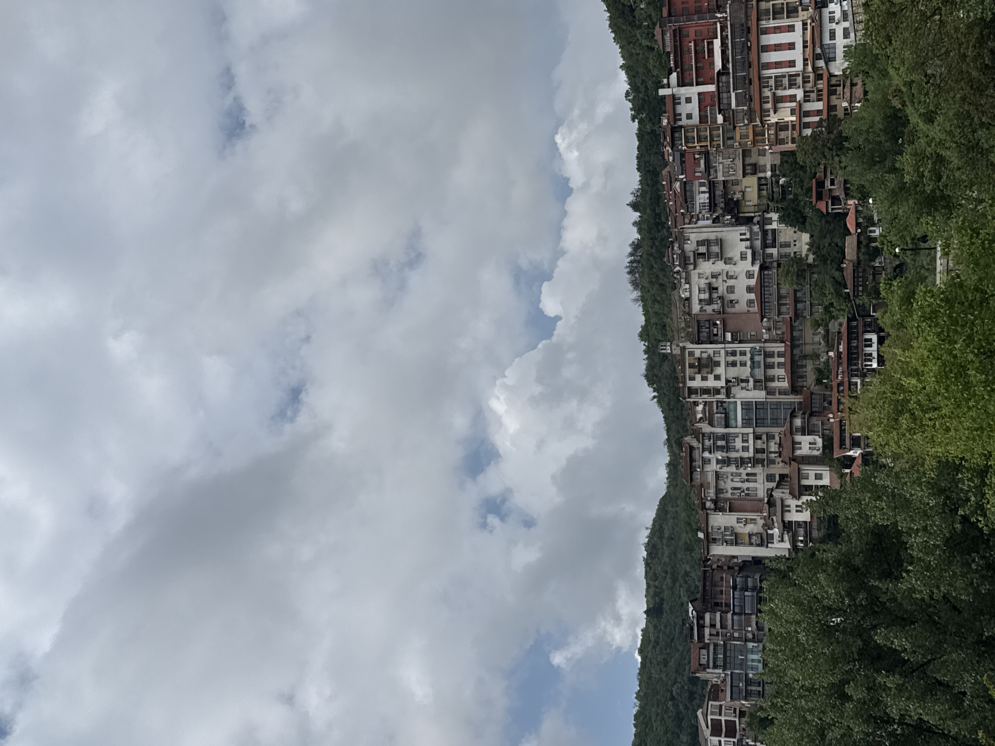 The image depicts a hillside in Veliko Tarnovo, Bulgaria, densely populated with buildings. The structures are multi-story, primarily white with red roofs, and appear to be residential. The hillside is covered in lush greenery, with trees visible in the foreground and interspersed amongst the buildings. The sky occupies a significant portion of the image, filled with white, puffy clouds against a pale blue backdrop. The overall perspective is slightly tilted.