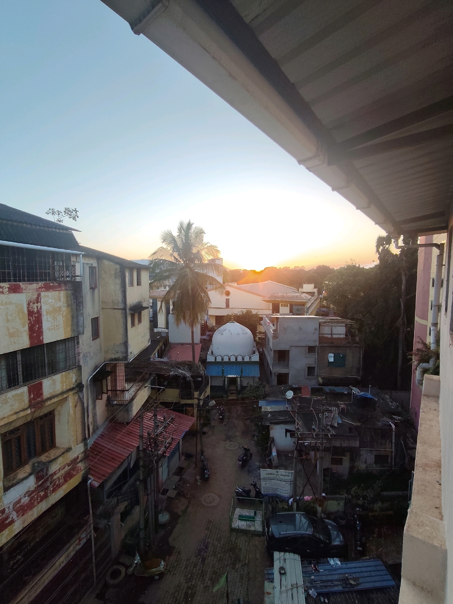 This image captures an urban street scene at dusk in Belagavi, India. The perspective is from a height, looking down onto a narrow street lined with buildings. A prominent, old, cream-colored building with red accents and barred windows stands on the left. Adjacent to it, a building with a red-tiled roof juts out over the street, supported by metal poles and electrical wires.
Further down the street, a white domed structure, possibly a mosque, is visible. Several smaller buildings with corrugated metal roofs are clustered together. A dark-colored car is parked in the street, and several scooters are scattered around. Trees, including a tall palm tree, are interspersed among the buildings.
The sky is a gradient of pale blue at the top, transitioning to a soft orange and yellow near the horizon, indicating either sunrise or sunset. The overall impression is of a densely populated, somewhat weathered urban environment. The image appears to have been taken from a balcony or window of a building overlooking the street. There are no visible people in the frame, but the presence of vehicles suggests ongoing activity.