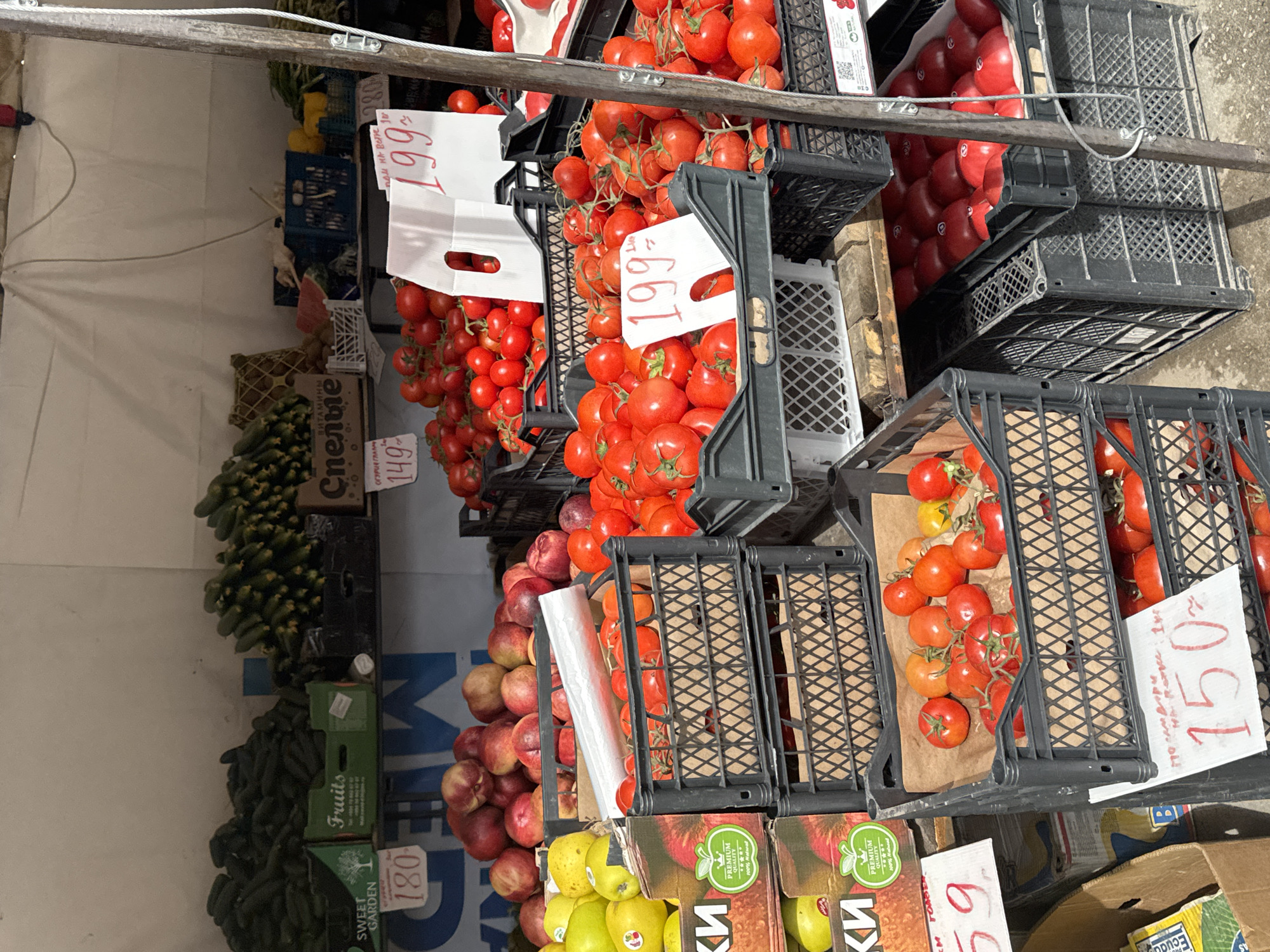 This image depicts a produce stand, likely located in Perm, Russia, based on Cyrillic text visible on price tags and packaging. The scene focuses on various fruits and vegetables arranged for sale, including tomatoes in black plastic crates priced at 199, 150 and 59 (currency not specified but likely Russian rubles), cucumbers in green boxes with labels such as "SWEET GARDEN" and "Fruits" priced at 149 and 180, and apples in boxes labeled "КИ". Price tags are handwritten. The stand appears to be covered by a white tarp. The scene includes crates stacked upon one another, creating a market display.