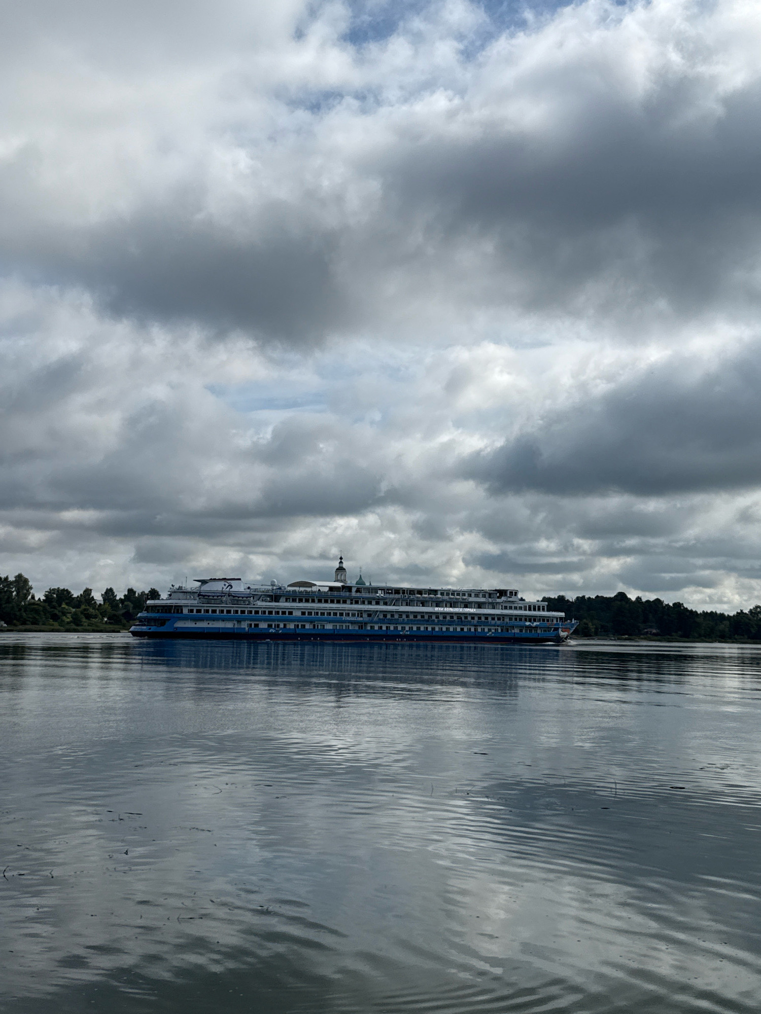 A long, multi-deck river cruise ship is navigating a wide, calm river under a cloudy sky. The ship, primarily white with blue accents and red trim along the waterline, has several rows of windows. A dark green treeline borders the river on the far bank. The water's surface is rippled and reflects the sky and ship. The location is the city of Rusanovo, Russia.