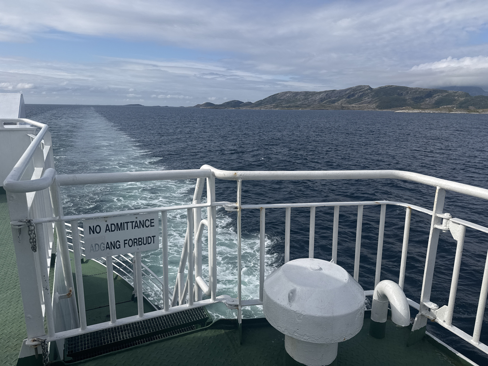 This image was taken from the deck of a ferry, looking towards the stern and out over the sea. The ferry is moving, as evidenced by the white wake trailing behind it. In the distance, the coastline of Onøya, Norway, is visible, characterized by rolling hills and mountains. The sky is partly cloudy, with a mix of blue sky and white clouds. On the deck, there is a white railing and a sign that reads "NO ADMITTANCE" and "ADGANG FORBUDT". A white, conical object, possibly a capstan or a vent, is also visible on the deck. The water is a deep blue, and the sea appears calm. The overall scene depicts a journey at sea with a view of the Norwegian landscape.