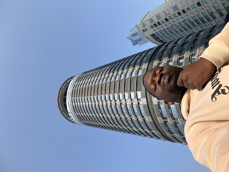 An adult male of dark complexion, with a beard, is depicted in a low-angle selfie outdoors on a clear day in Yuzhong District, Chongqing, China. He wears a light-colored hooded sweatshirt with a dark graphic partially visible on the chest. His right hand is positioned beneath his chin, and he looks directly into the camera with a neutral expression. In the background, a tall, cylindrical skyscraper dominates the frame, identified as the JW Marriott Hotel Chongqing due to the visible "JW" and Chinese characters ("万豪") on its upper facade. To the right of this primary building, another high-rise structure with a lighter-colored facade and a distinctive angular glass top section is partially visible against a clear blue sky.