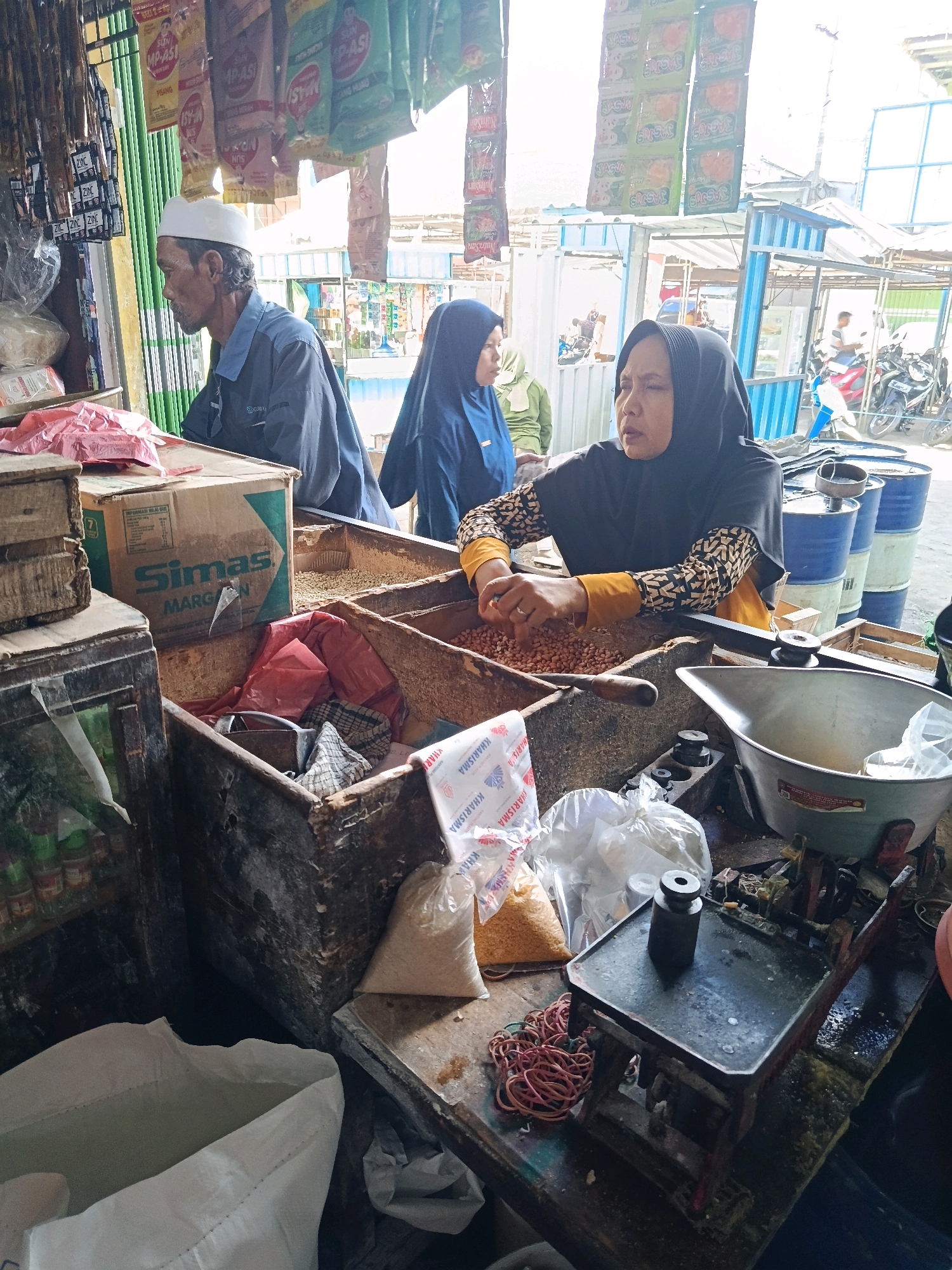 The image depicts a market stall in Senduro, Indonesia, with an assortment of goods and people. In the foreground, a woman wearing a black hijab and an orange and black patterned shirt is actively sorting peanuts in a wooden bin. To her left, a man in a blue shirt and white skullcap is looking away from the camera, his hands resting on a box labeled "Simas Margarin." Behind them, another person in a blue hijab is partially visible.
The stall is filled with various items including bags of what appears to be grains or seeds, plastic bags of goods, and stacks of barrels in the background. Packaged food items, some with colorful labels, hang from the ceiling. A metal scale with weights and a bundle of red rubber bands are prominent on the counter in the lower right portion of the image. The overall environment suggests a bustling local marketplace with a focus on food products.