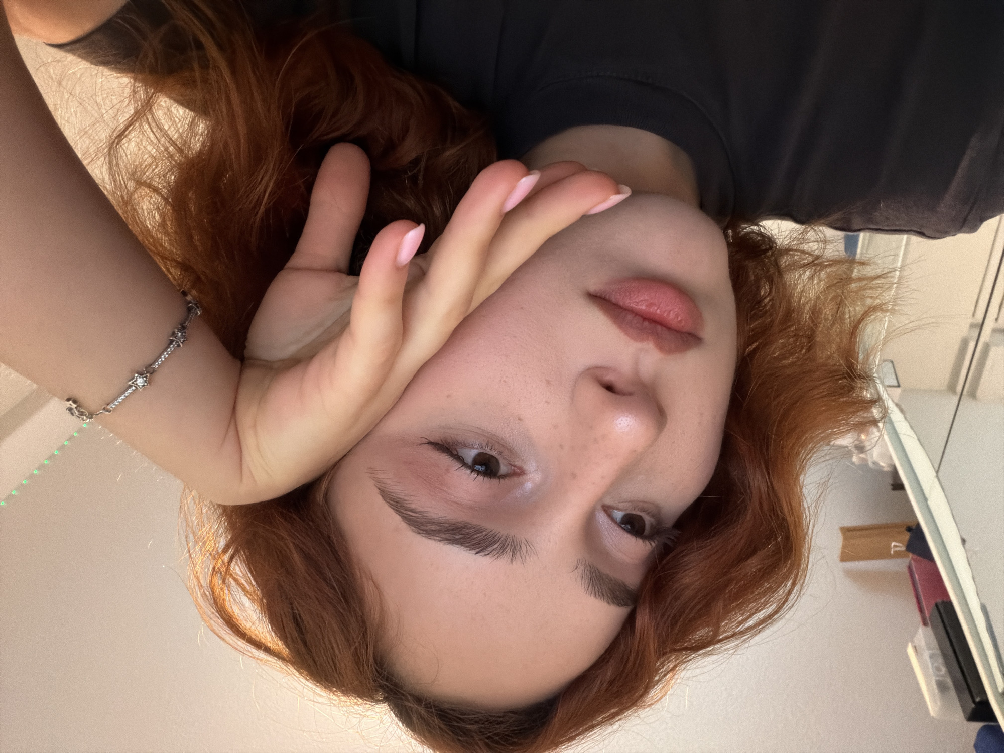 The image is a close-up, angled shot of a young woman with auburn hair, resting her head on her hand. She is wearing a dark t-shirt and a bracelet on her wrist. Her makeup includes pink eyeshadow and lip color. The background appears to be an indoor setting, with a shelf visible on the left containing some objects. There is no indication of the city of Strasbourg, France in the image.