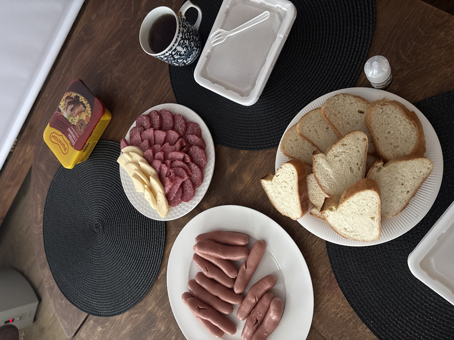 An overhead view captures a dining setup on a dark brown wooden table in Lezhnevo, Russia. The table is adorned with several black, circular, woven placemats. Food items are arranged on white plates: one plate contains approximately twelve pinkish sausages, some exhibiting longitudinal cuts; another plate displays numerous slices of red salami alongside crescent-shaped slices of pale yellow cheese; a third plate holds an assortment of sliced white bread. A yellow rectangular package, labeled "Роллтон" (Rollton) "Лапша с говядиной" (Noodles with beef), rests on a placemat. A blue and white patterned ceramic mug contains a dark liquid. Adjacent to the mug is a white rectangular disposable foam container with a clear plastic fork placed inside. A small, clear-topped shaker is also visible on the table. A grey electronic device is partially visible in the upper left background, and a white horizontal surface is in the upper right background. No individuals are present in the image.
