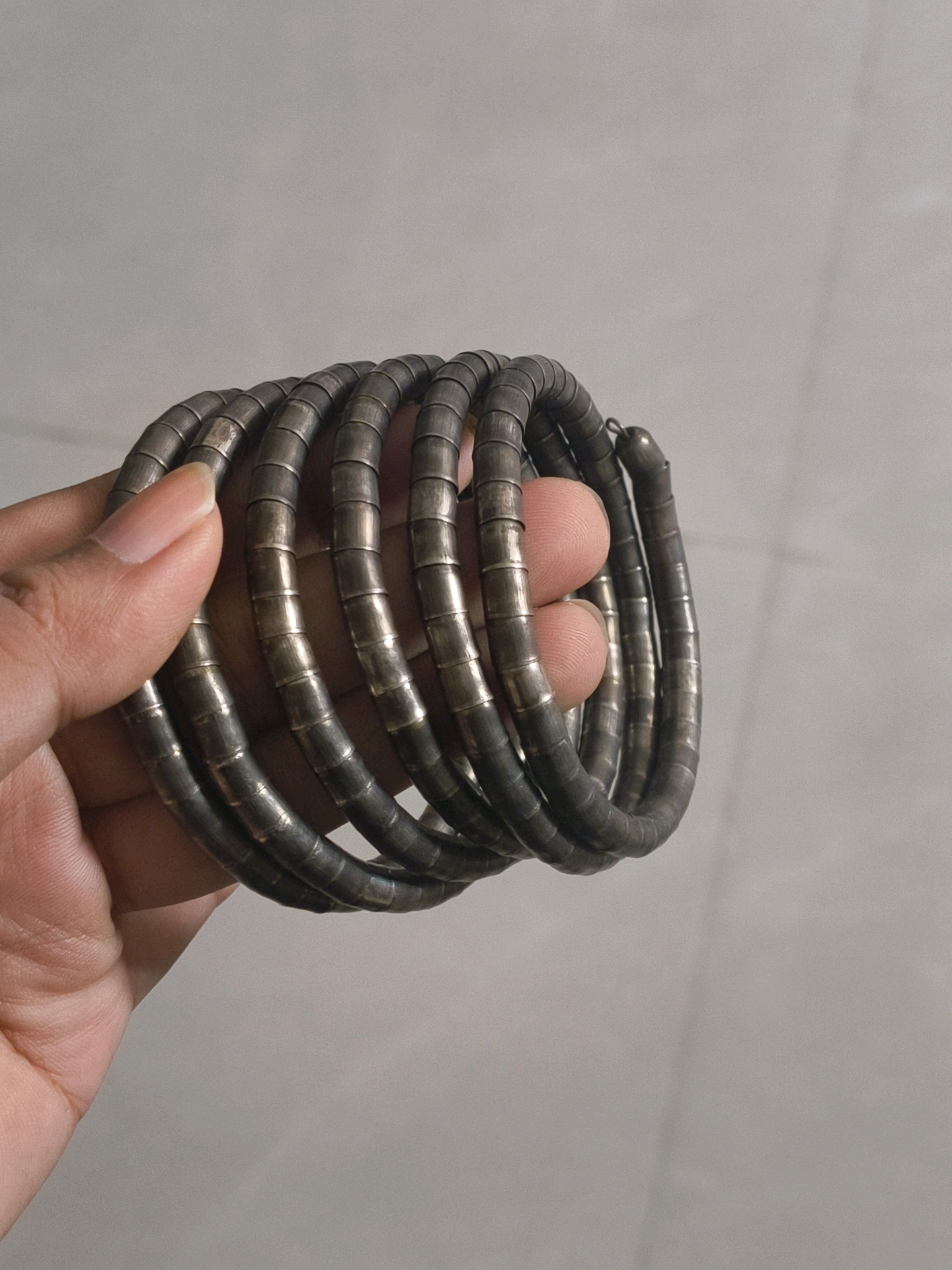 An overhead shot shows a hand in the foreground holding a coiled, metallic bracelet. The bracelet is comprised of multiple tightly wound segments that resemble a spring or a segmented tube. The hand's skin tone is light brown, with visible fingerprints and the texture of the skin. The background consists of a plain, light gray wall. The image captures the texture and metallic sheen of the bracelet, as well as the details of the hand. This scene is located in Mohali, India.