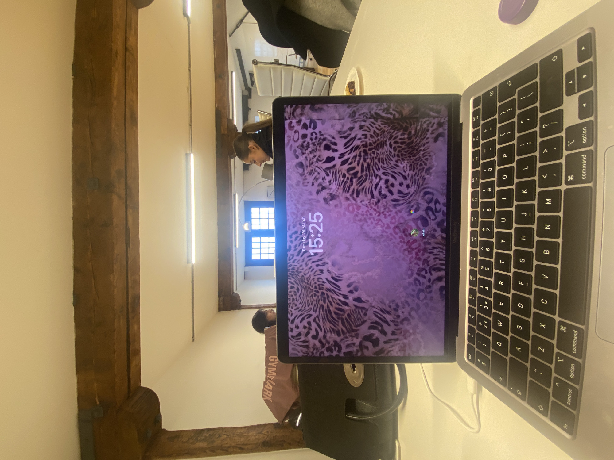 The image depicts an indoor office or co-working space within the city of London, United Kingdom, characterized by exposed, rough-hewn wooden ceiling beams. The immediate foreground features an open silver Apple MacBook laptop, displaying a lock screen or wallpaper with a pink and purple leopard print pattern. The screen clearly shows "Tuesday 24 March" and the time "15:25", with "admin" and a small profile icon visible at the bottom right. A white charging cable is connected to the laptop's left side. The laptop's keyboard has a QWERTY layout, including a '€' symbol on the '3' key. To the right of the laptop, a purple cylindrical object rests on the white desk surface.
In the midground, two individuals are seated at separate workstations behind the laptop. On the left, a person with dark hair is partially visible behind a black handbag with a silver circular clasp. A brown/maroon Gymshark branded bag, with the logo inverted, is draped over the black bag. On the right, a woman with dark hair pulled back in a ponytail is seen in profile, facing left. She is seated at a white desk, and a white modern office chair with a ribbed back is positioned behind her. A small white plate containing a dark-colored pastry or food item is on the desk to her right.
The background reveals white walls and ceilings with multiple fluorescent strip lights. Two windows with dark, multi-paned frames are visible, allowing natural light into the space. The architectural elements suggest an older building converted into a modern workspace, consistent with commercial properties found within London.