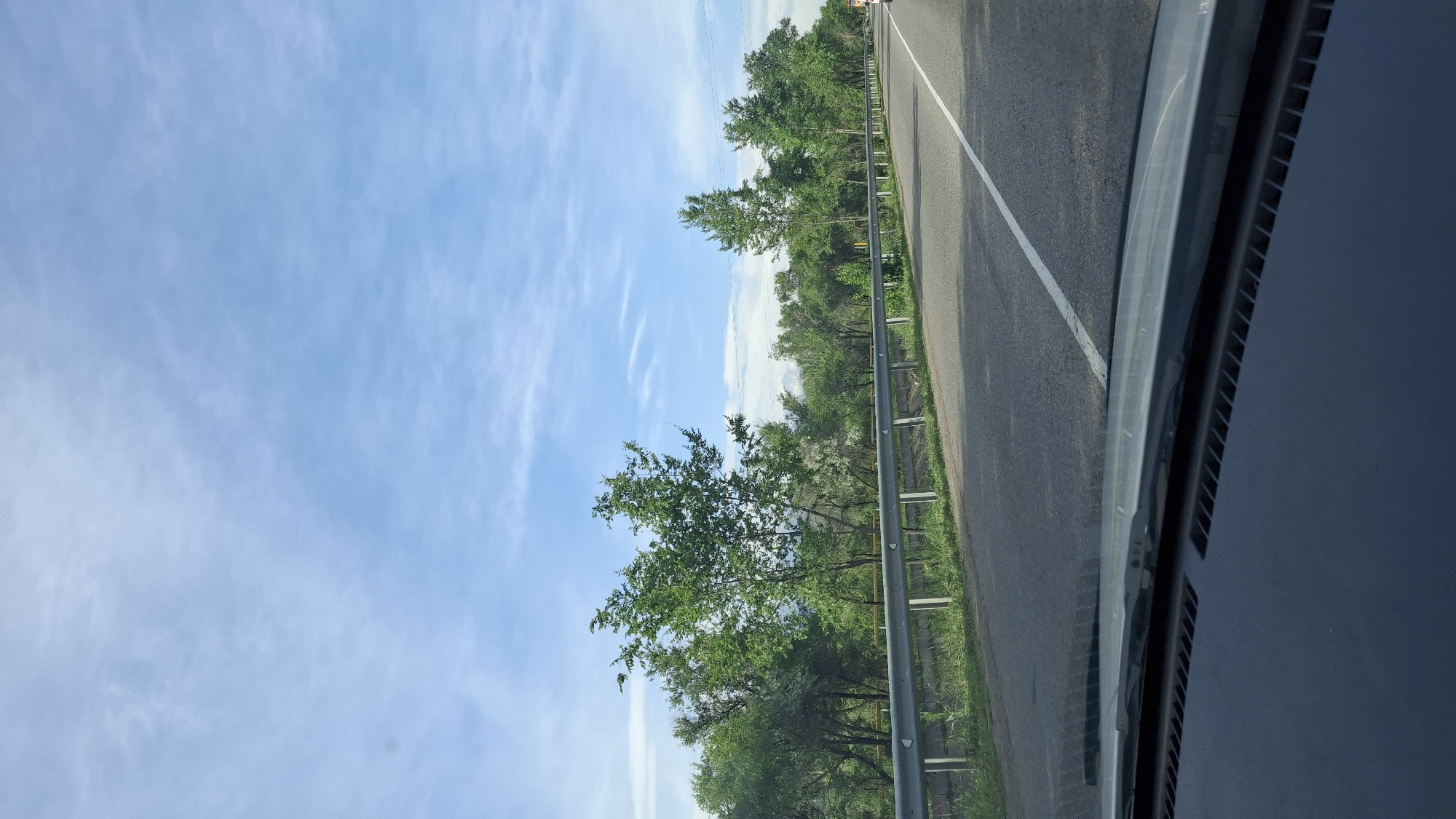 The image shows a view from inside a vehicle, likely a car, while traveling on a road in Түзусай, Kazakhstan. The road has a white line marking, and a metal guardrail is present along the side. Beyond the guardrail, there are trees and greenery. The sky is visible above the trees, with light clouds scattered across a blue background. The dashboard and hood of the car are visible in the lower right portion of the image.