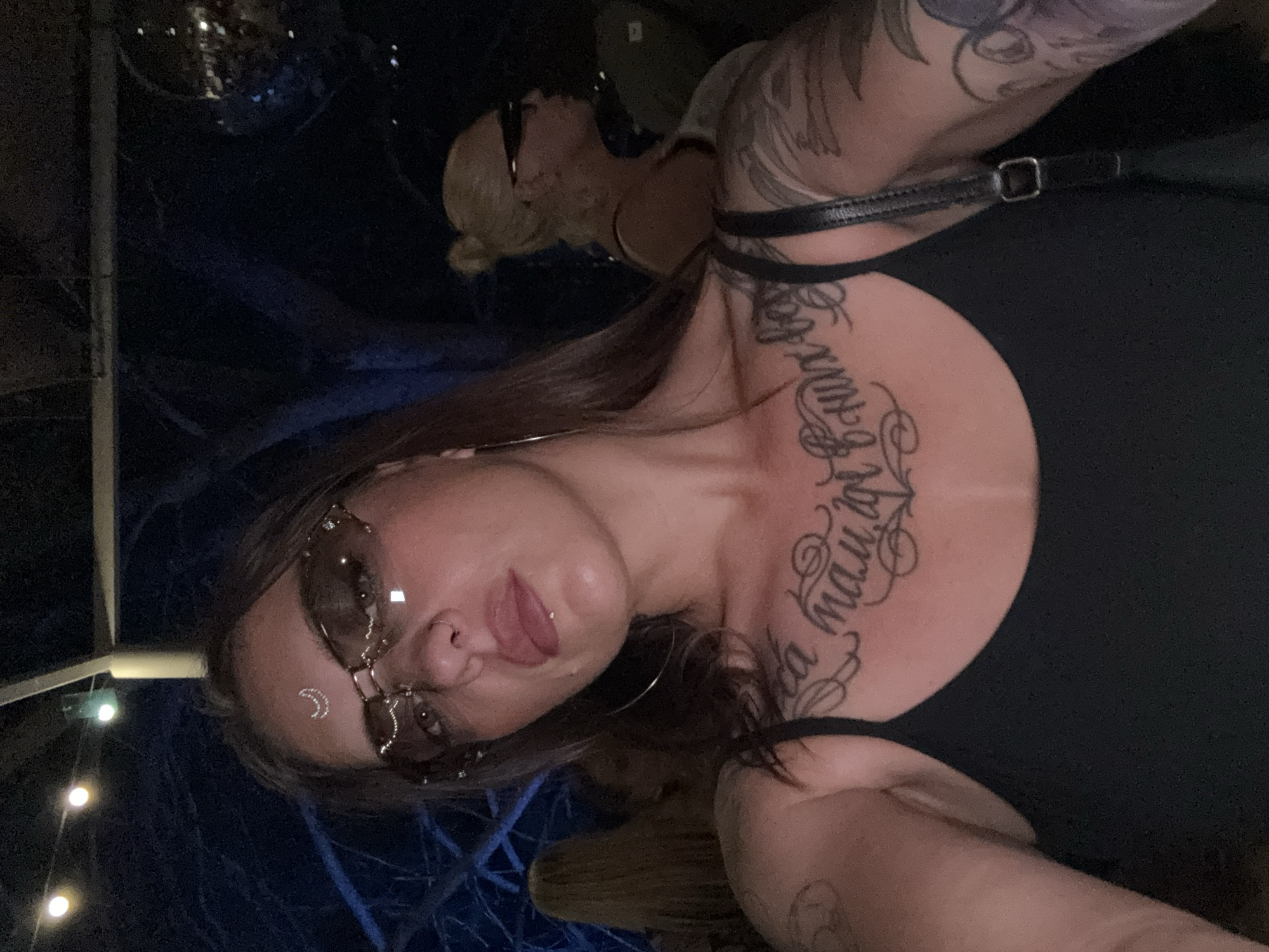 This is a close-up, selfie-style portrait of a woman indoors at night. She is wearing sunglasses with rhinestones on them and a moon-shaped rhinestone decoration on her forehead. She has a nose ring and a lip ring. Her hair is long and brown, and she is wearing a black tank top. She has extensive tattoos on her chest and arms. In the background, other people are visible, as well as some string lights and what appears to be a disco ball. The overall lighting is dim and atmospheric. The setting is likely a club or a bar, possibly in the city of Sochi, Russia, given the provided context of location.