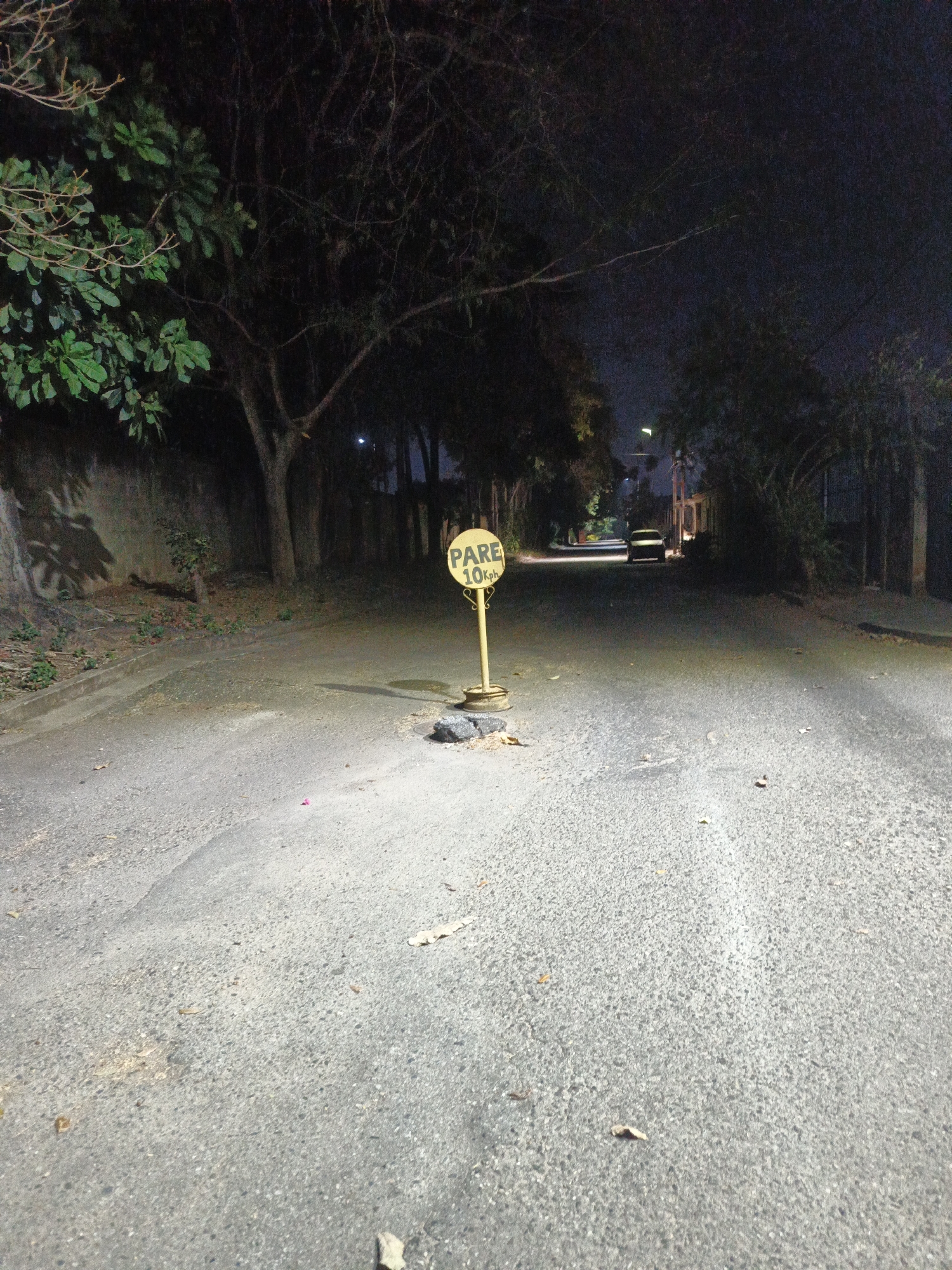 The image depicts a dimly lit street in Tinaquillo, Venezuela, at night. In the foreground is an asphalt road surface littered with debris. A yellow "PARE 10Kph" (STOP 10Kph) sign is positioned in the center, with damaged pavement surrounding its base. Trees border the street on both sides, casting shadows. In the distance, a car is visible parked on the street, its headlights illuminating the scene. The overall environment suggests a residential area.