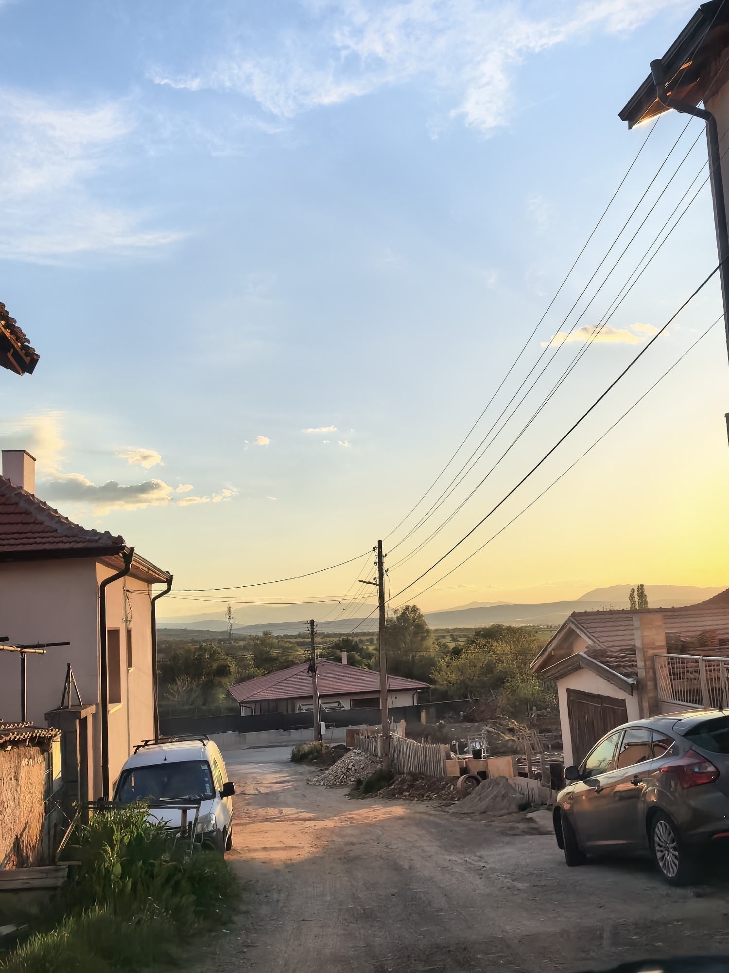 The image depicts a dirt road in Sapareva Banya, Bulgaria, lined with buildings and vehicles. A white van is parked on the left, obscured by greenery. A dark-colored car is parked on the right. Construction materials, including piles of dirt and wooden structures, are visible between the vehicles. Power lines stretch across the sky. In the background, mountains are visible against a sky transitioning from blue to a yellow sunset hue. The overall scene suggests a residential area undergoing construction or renovation.