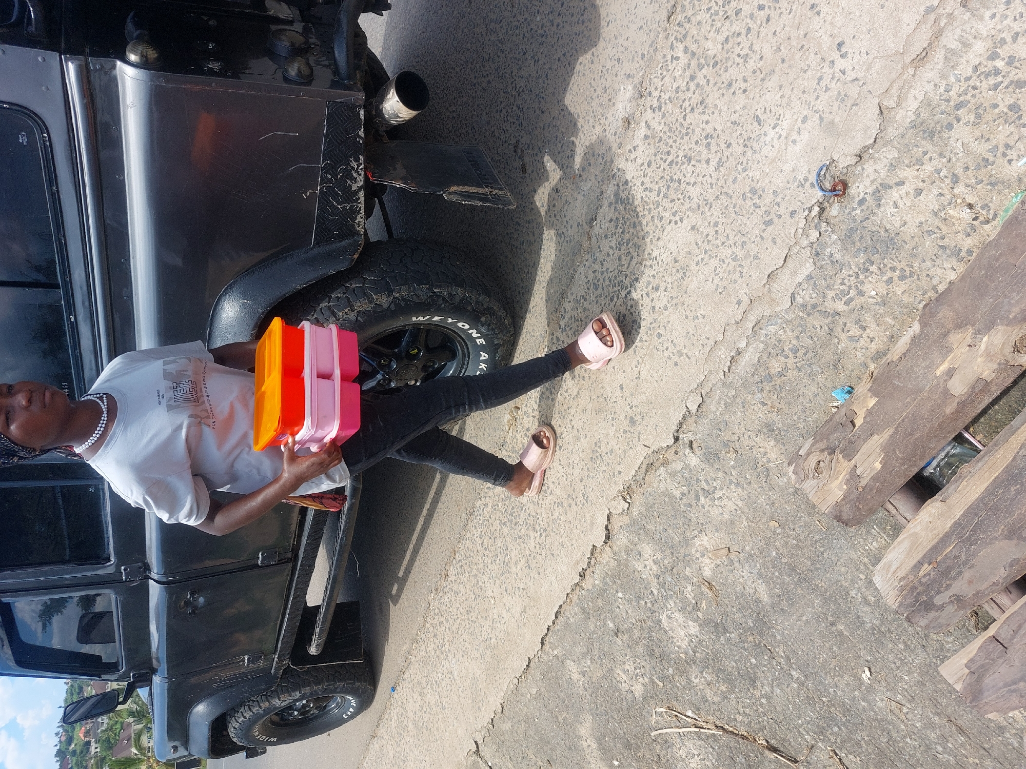 A young adult female is depicted walking rightward on a paved road, carrying three stacked plastic food containers (one orange, two pink) in her left hand. She wears a light grey t-shirt, dark trousers, a patterned headscarf, a white beaded necklace, and light pink slide sandals. To her right, a dark-colored Land Rover Defender 110 is stationary or moving slowly, partially obscuring the background. The vehicle exhibits off-road tires, running boards, and signs of wear, including scratches and dirt on its bodywork; its rear features a mud flap and a chrome exhaust pipe. The road surface is asphalt, displaying cracks and aggregate. In the foreground, on a lower concrete section, three weathered wooden planks are present alongside dry foliage and a small rusty metal ring with a blue cord. The background reveals a hilly terrain densely populated with residential buildings nestled among green vegetation under a partly cloudy sky, consistent with an elevated residential area in Dar es Salaam, Tanzania.
