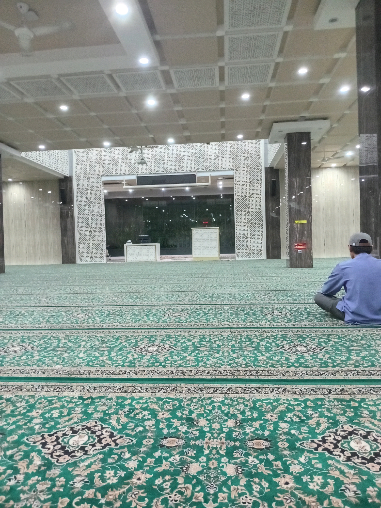 The image depicts the interior of a mosque, specifically the prayer hall. The floor is covered with a large, green carpet featuring intricate patterns. In the foreground, a person wearing a blue shirt and a grey cap is seen sitting cross-legged on the carpet, facing away from the camera towards the front of the mosque. The mosque's architecture is modern, with a patterned ceiling, recessed lighting, and decorative panels on the walls. There is a raised platform with a lectern and a screen at the front, suggesting an area for prayer or religious services. The overall environment appears clean and spacious, characteristic of a place of worship. The scene is set in Randuagung, Indonesia.