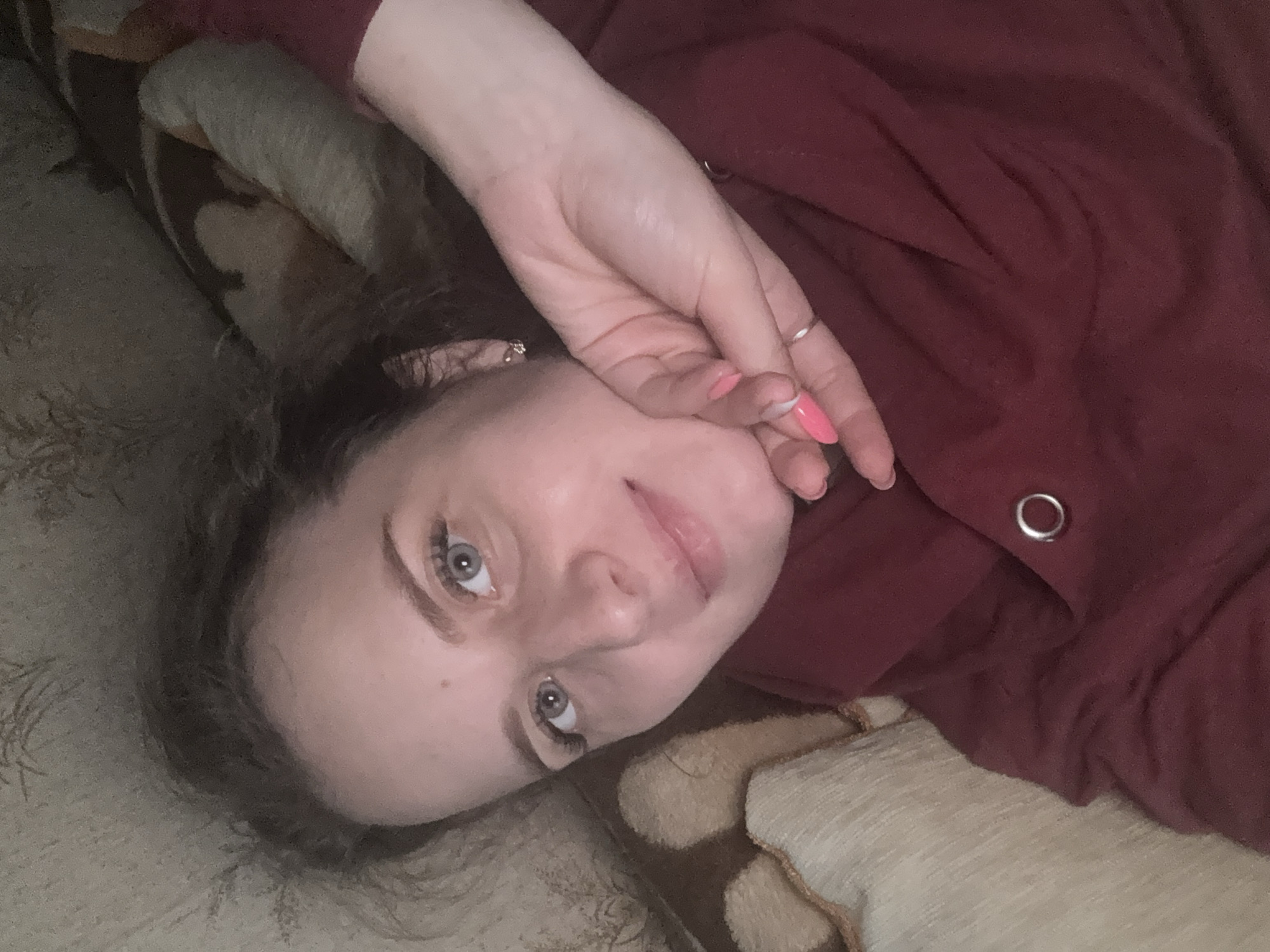 The media file is a close-up, over-the-shoulder selfie of a young woman lying down. She is wearing a dark red, long-sleeved garment with a metal grommet detail on the sleeve. Her hair is dark and appears to be loosely curled, falling around her face and shoulders. Her right hand is raised, partially covering her face, with manicured nails painted bright pink. She is wearing a ring on her ring finger.
The woman has light skin, light blue eyes, and is looking towards the camera. Her facial expression is neutral or slightly contemplative. The background consists of a textured surface, possibly a couch or bed, with a light beige and brown patterned fabric. The lighting is soft and appears to be indoor, suggesting it is either daytime or evening. The image is oriented sideways, as if taken while the subject is lying on their side. No text is visible in the image. The location context of Severnyy, Russia, does not provide any specific visual cues within the image itself.
