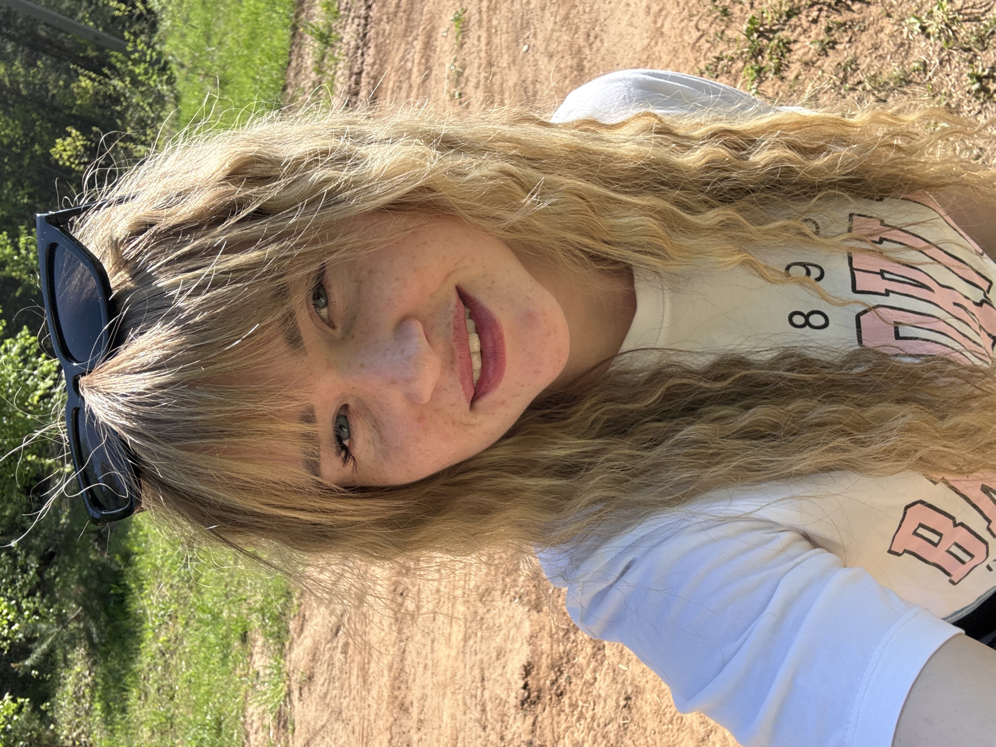 The image depicts a blonde woman with fair skin and freckles, likely in her late teens or early twenties. She is wearing a white t-shirt with a graphic print and has a pair of black sunglasses perched atop her head. The woman is smiling. The setting appears to be a dirt path or road with green foliage visible in the background, possibly located in a rural or semi-rural area near Марковский, Russia. The environment suggests an outdoor location, possibly a park or wooded area.