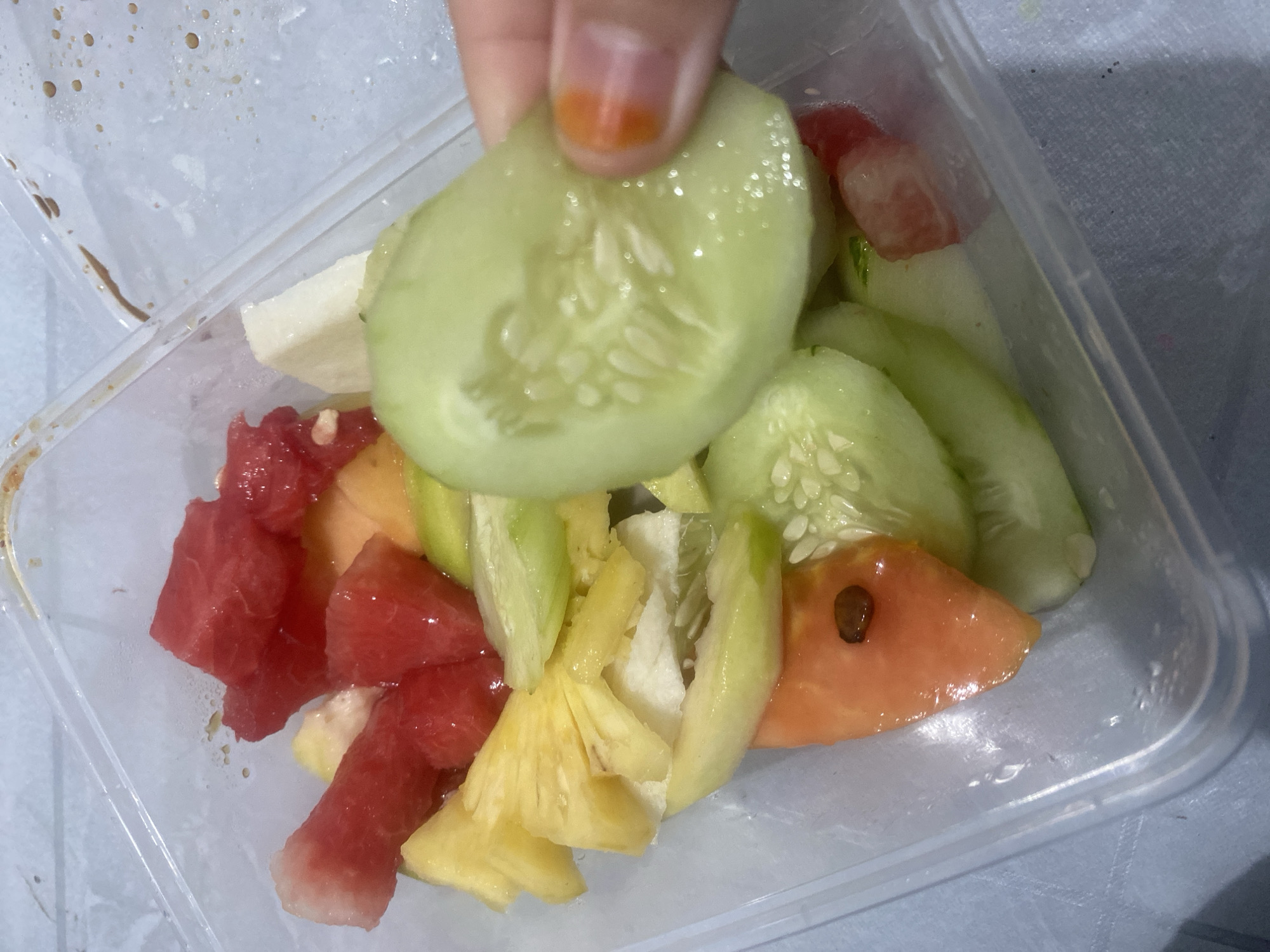 A clear plastic container filled with a variety of chopped fruits and vegetables is shown from a top-down perspective. The contents include vibrant red cubes of watermelon, pale yellow chunks of pineapple, and slices of what appears to be cantaloupe or honeydew melon. Interspersed among the fruits are several slices of cucumber, one of which is being held by a person's fingers, revealing the seedy interior. A few pieces of tomato are also visible, one with a prominent dark seed. The container is placed on a textured, light-colored surface that resembles a table or countertop. A hand with a fingernail painted orange is visible at the top of the frame, holding a slice of cucumber. The overall impression is of a fresh, prepared salad or fruit mix, likely in a home or casual dining setting. There are small water droplets and some light brown smudges on the inside and outside of the plastic container, suggesting it might have been recently prepared or handled. The lighting appears to be artificial, with some highlights reflecting off the moist surfaces of the produce and the plastic. No visible text is present in the image.