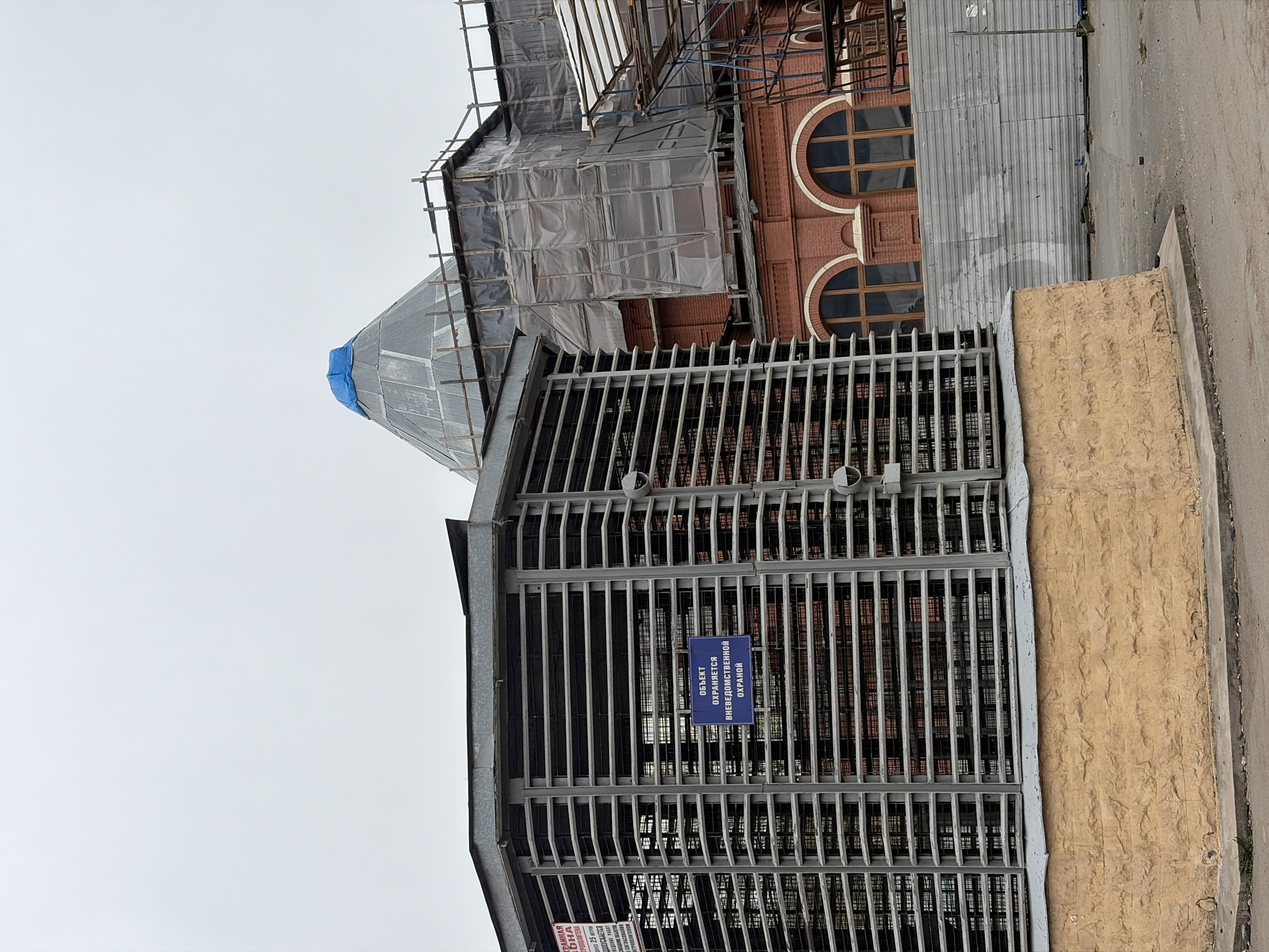 Here's a description of the image:
The image captures a building undergoing construction in what appears to be Saint Petersburg, Russia. Scaffolding and protective coverings obscure part of the building's facade, particularly around the upper levels, which include a tower-like structure. The building features a red brick exterior with arched windows. A metal grate encloses the base of the building or an adjacent area. A blue sign attached to the grate displays text in Russian indicating that the property is protected by non-departmental security. The overall impression is one of ongoing renovation or restoration.