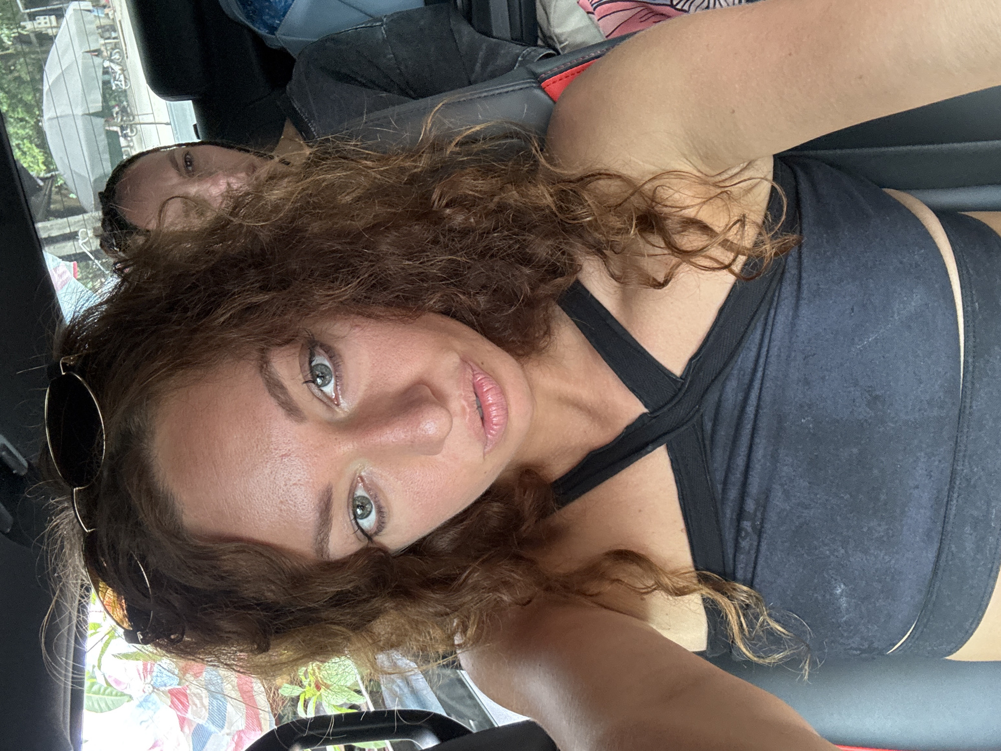 The image shows a woman with curly brown hair and green eyes taking a selfie from inside a vehicle. She is wearing a black halter-style top. Behind her, another person with dark hair is visible in the passenger seat. Through the rear window, a street scene with parked motorcycles and an umbrella is visible, suggesting an outdoor environment in a city. The backdrop appears to be the city of Bắc Yên, Vietnam.