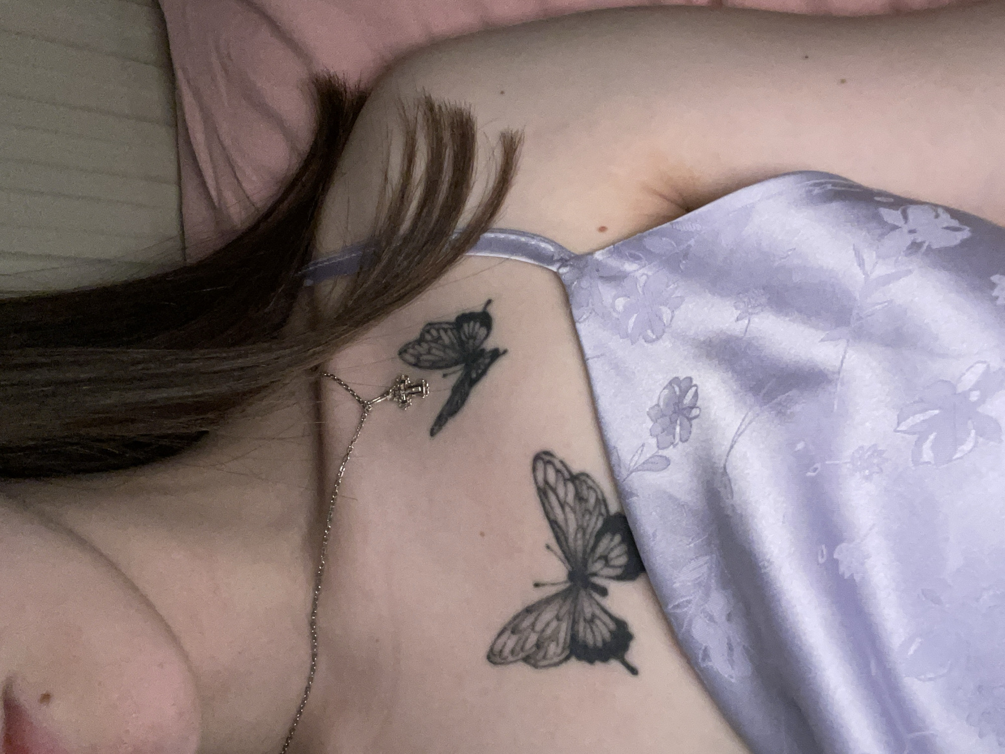 A close-up, angled view of a person lying down. They have light skin and brown hair. They are wearing a lilac silk camisole with a floral pattern. Two butterfly tattoos are visible on their chest and shoulder area. A silver necklace with a small charm is visible on their chest. A pink pillow is partially visible in the background. The image does not provide information related to the city of Chaikovsky, Russia.