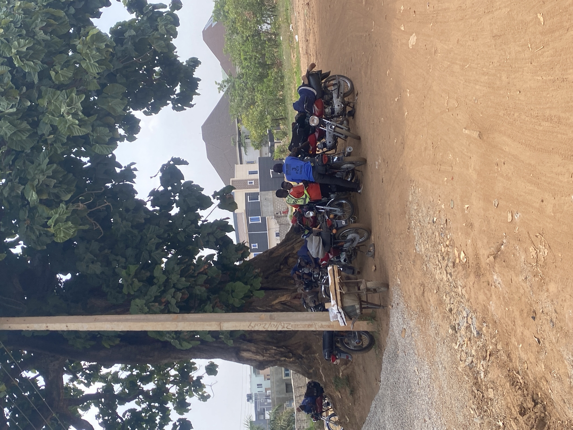 The image captures an outdoor scene, likely in a suburban or semi-urban area of Abuja, Nigeria. A large, mature tree with dense green foliage dominates the left side of the frame, providing shade over a gathering of people and motorcycles.
In the foreground, a dusty, reddish-brown dirt road or open ground is visible, with patches of gravel. Under the tree, a line of at least seven motorcycles is parked. Several men are standing or gathered around these bikes. One man in the center wears a blue shirt with "FCT SOCIAL" clearly written on the back. Another person nearby is seen wearing a green and yellow reflective vest. Further down the line, a person is lying across the seat of a red motorcycle, seemingly resting. An old wooden cart with a container sits between some of the bikes.
A tall, rectangular concrete utility pole is positioned near the tree's trunk. Visible text on the pole includes the date "25/10/2019" and some other illegible markings. In the background, beyond the motorcycles and tree, residential buildings with distinct roofs are visible against an overcast or hazy sky. The scene suggests a common waiting or gathering spot, possibly for motorcycle taxi operators, under the shelter of the tree during the day. The lighting is diffused, indicating either an overcast day or early morning/late afternoon.