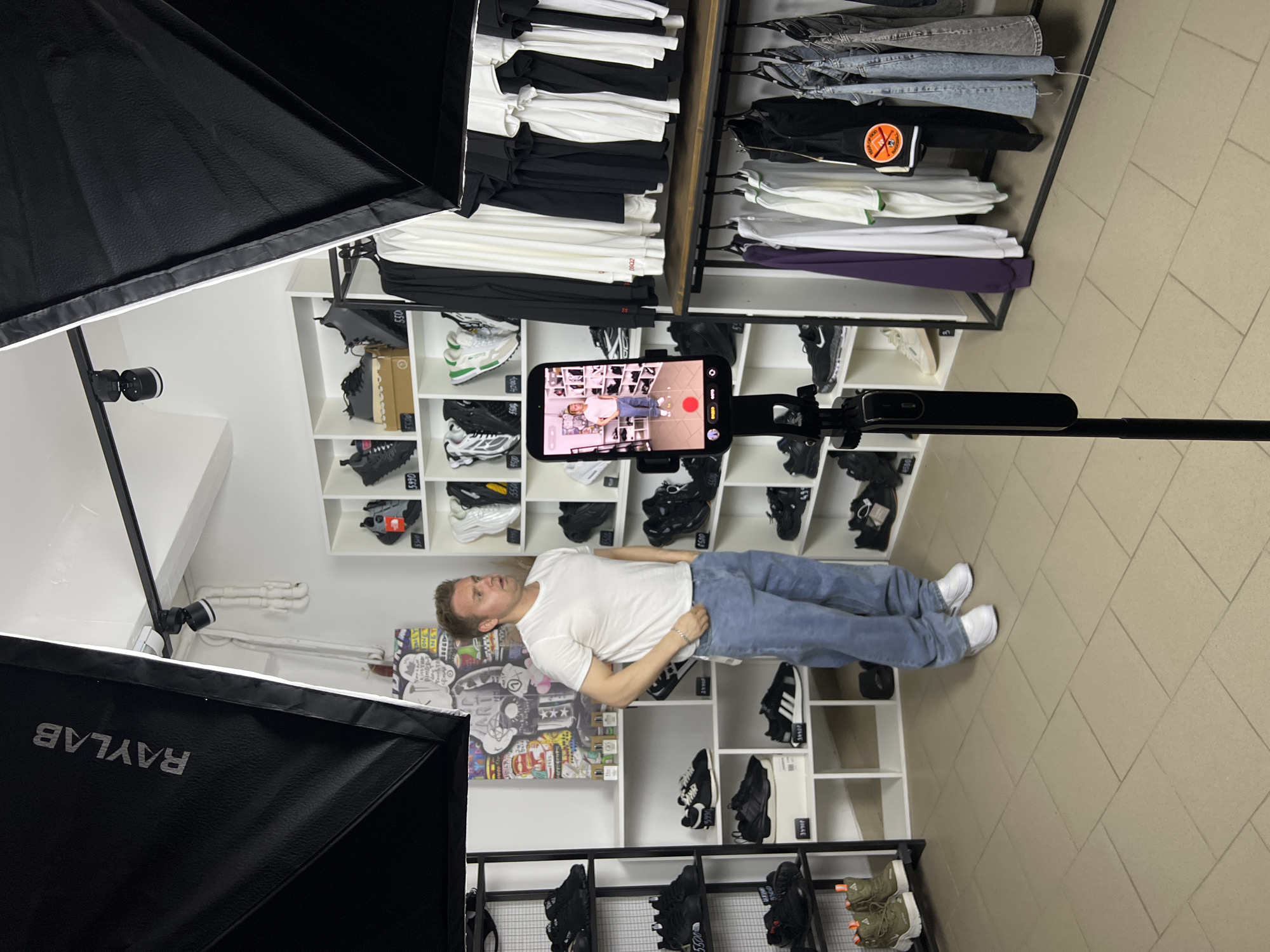 This image, likely taken in Chaikovsky, Russia, depicts an indoor retail space or studio setup where a man is recording content. The scene is primarily illuminated by two large, black softbox lights, one of which prominently displays the brand name "RAYLAB" in white text.
In the center of the frame, a man wearing a white t-shirt, light blue jeans, and white sneakers stands in front of several white shelves. These shelves are neatly stocked with various sneakers, including black, white, and some with green accents, and folded apparel such as white, black, and purple t-shirts or sweatshirts. Some price tags are visible on the shelves, showing numbers like "5300" and "5990". Behind the man, on one of the shelves, there is a piece of colorful, graffiti-style artwork.
The man is looking towards a smartphone mounted on a black tripod or selfie stick, positioned directly in front of him. The phone's screen is visible, showing a live camera view of the man, indicating he is either filming himself or being filmed. The phone displays typical camera interface elements, including a red record button, a timer, and options for "PHOTO" and "VIDEO." The overall setup suggests a professional or semi-professional recording session, possibly for social media content or a product review, within what appears to be a fashion or sneaker store.